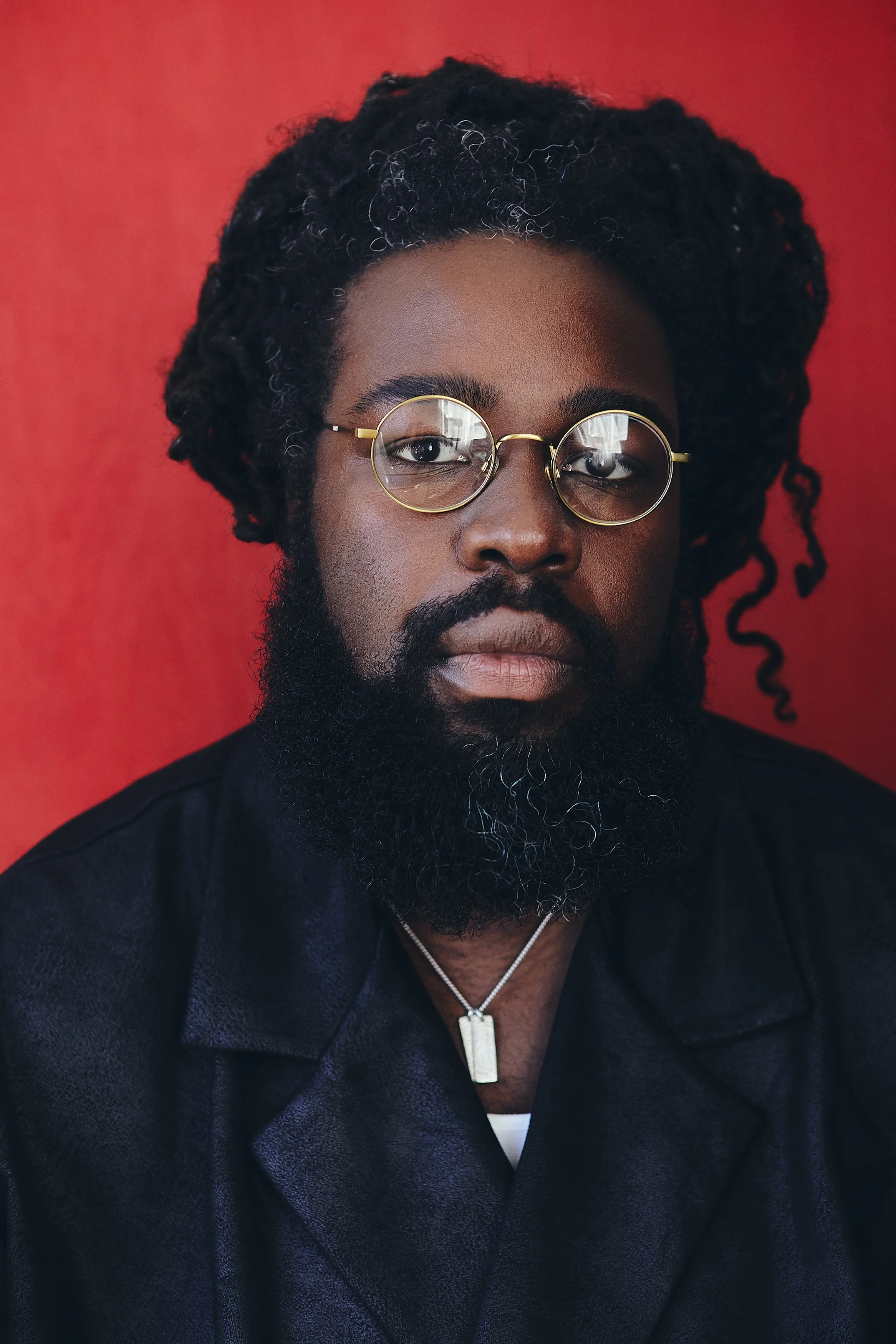 A man with dark skin, thick curly hair, a beard, and glasses, wearing a dark jacket and a silver necklace, standing against a red background.