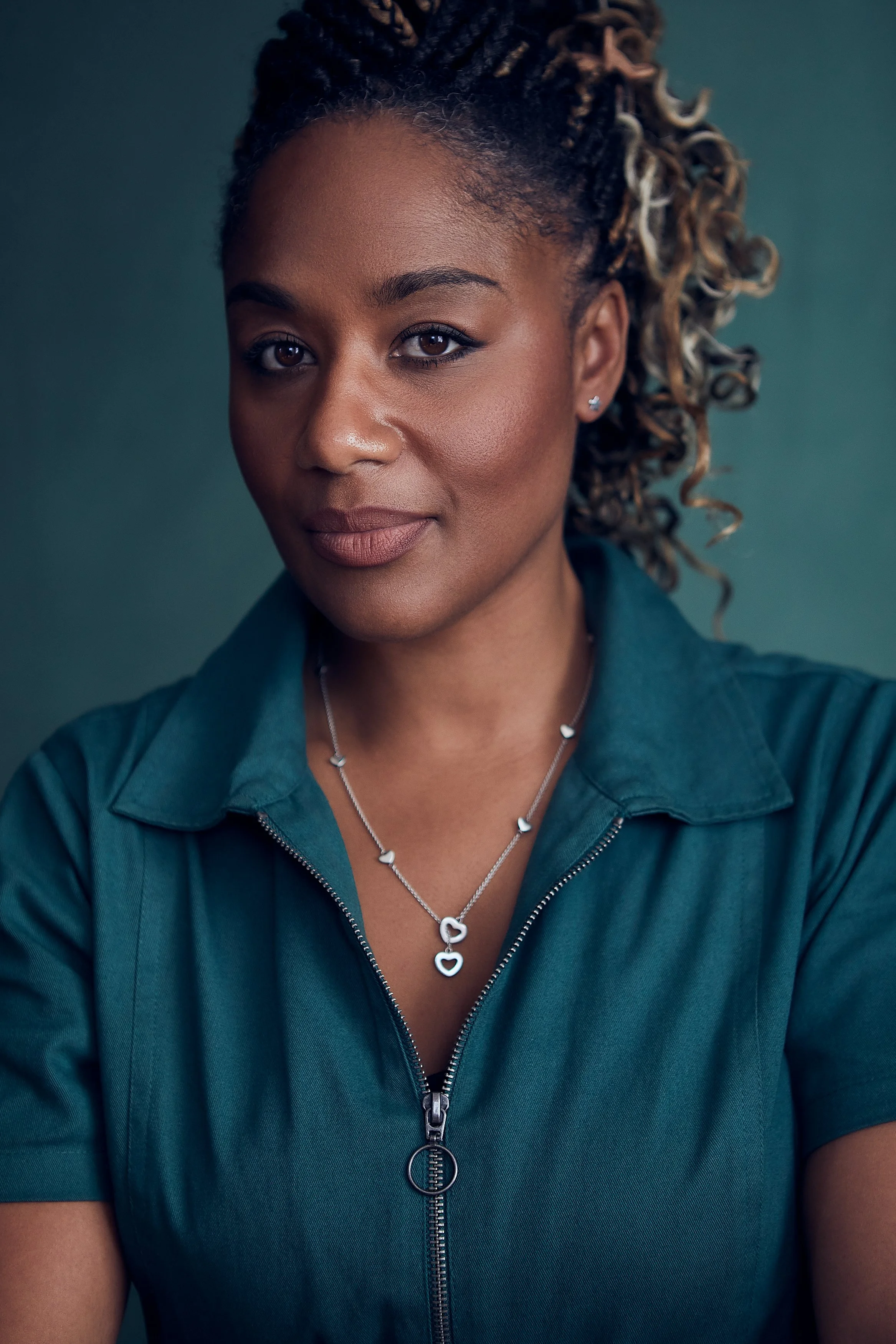 A portrait of a woman with curly, styled hair wearing a teal zip-up shirt and a silver necklace with heart-shaped charms.