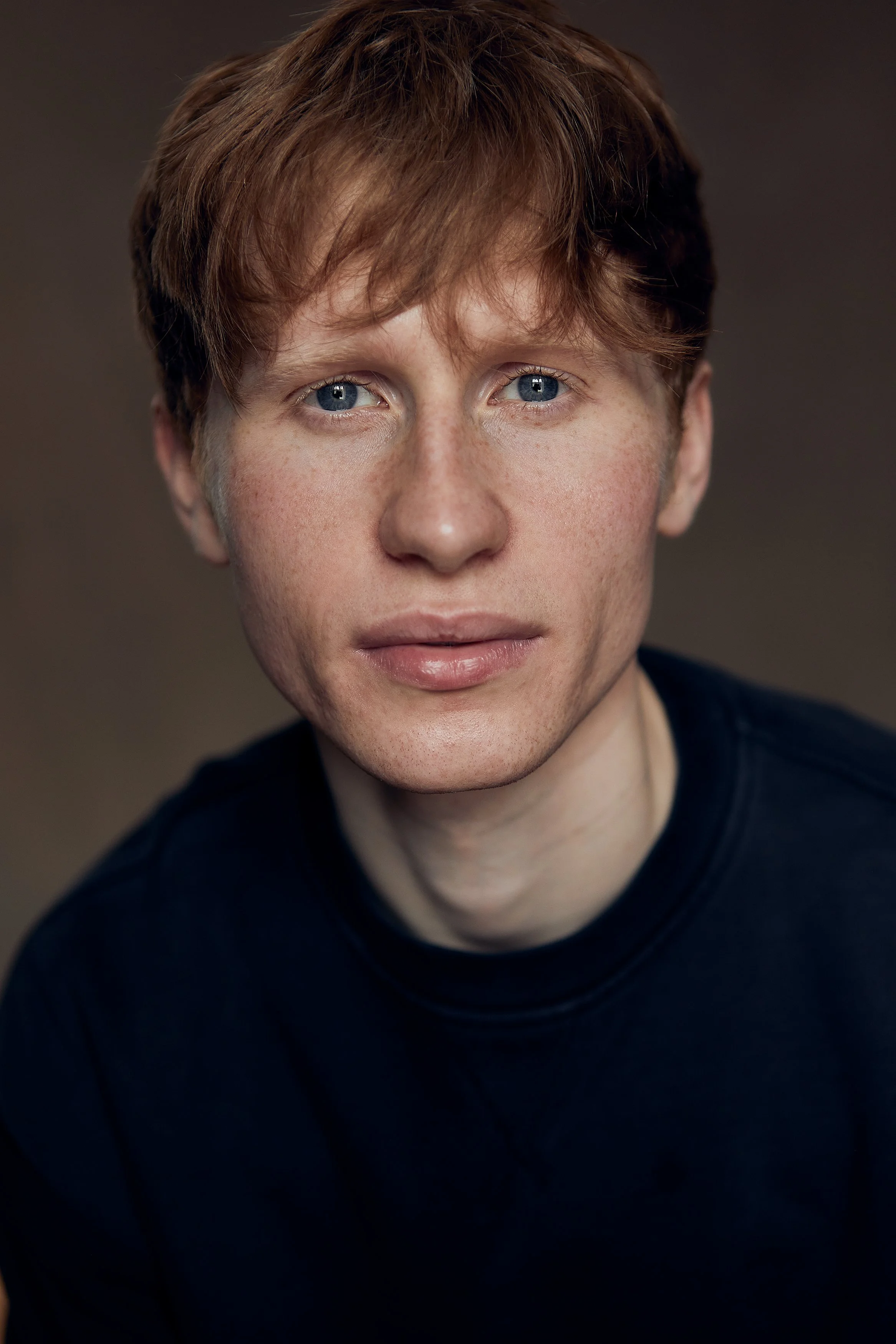 Close-up of a young man with ginger hair, blue eyes, and fair skin, wearing a black shirt against a plain background.