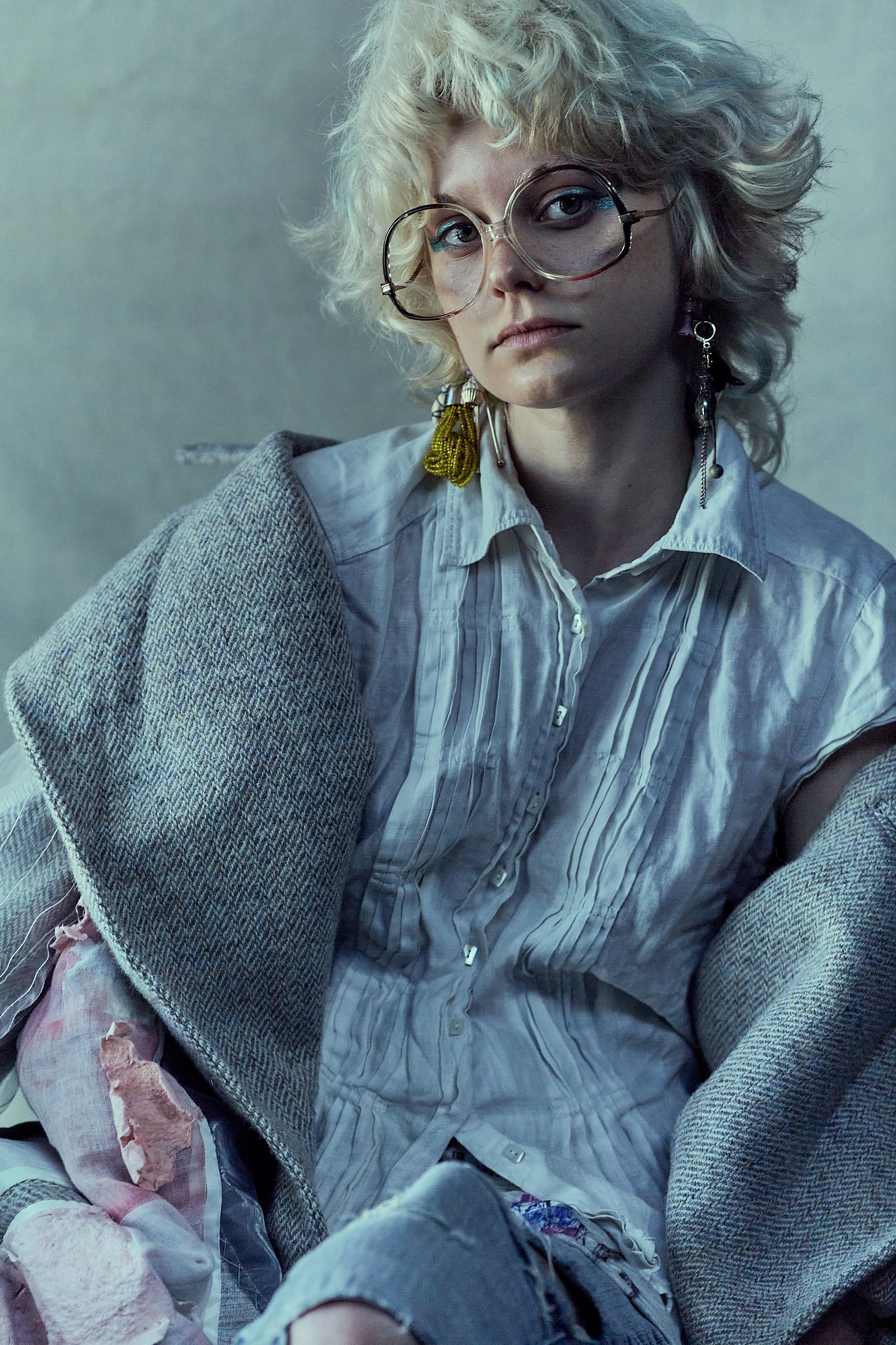 A person with curly blonde hair, wearing big round glasses, a light-colored striped shirt, and layered clothing, looking directly at the camera.