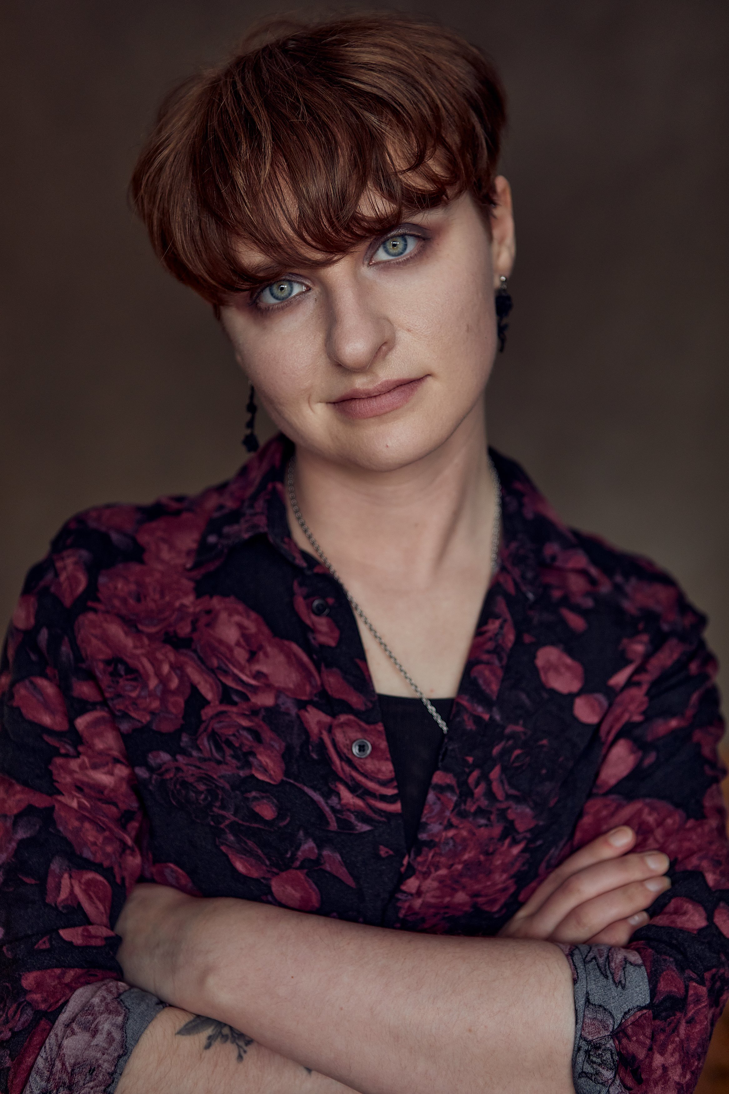A woman with short, reddish-brown hair and light blue eyes, wearing a dark floral shirt, earrings, and a necklace, with arms crossed against a dark background.