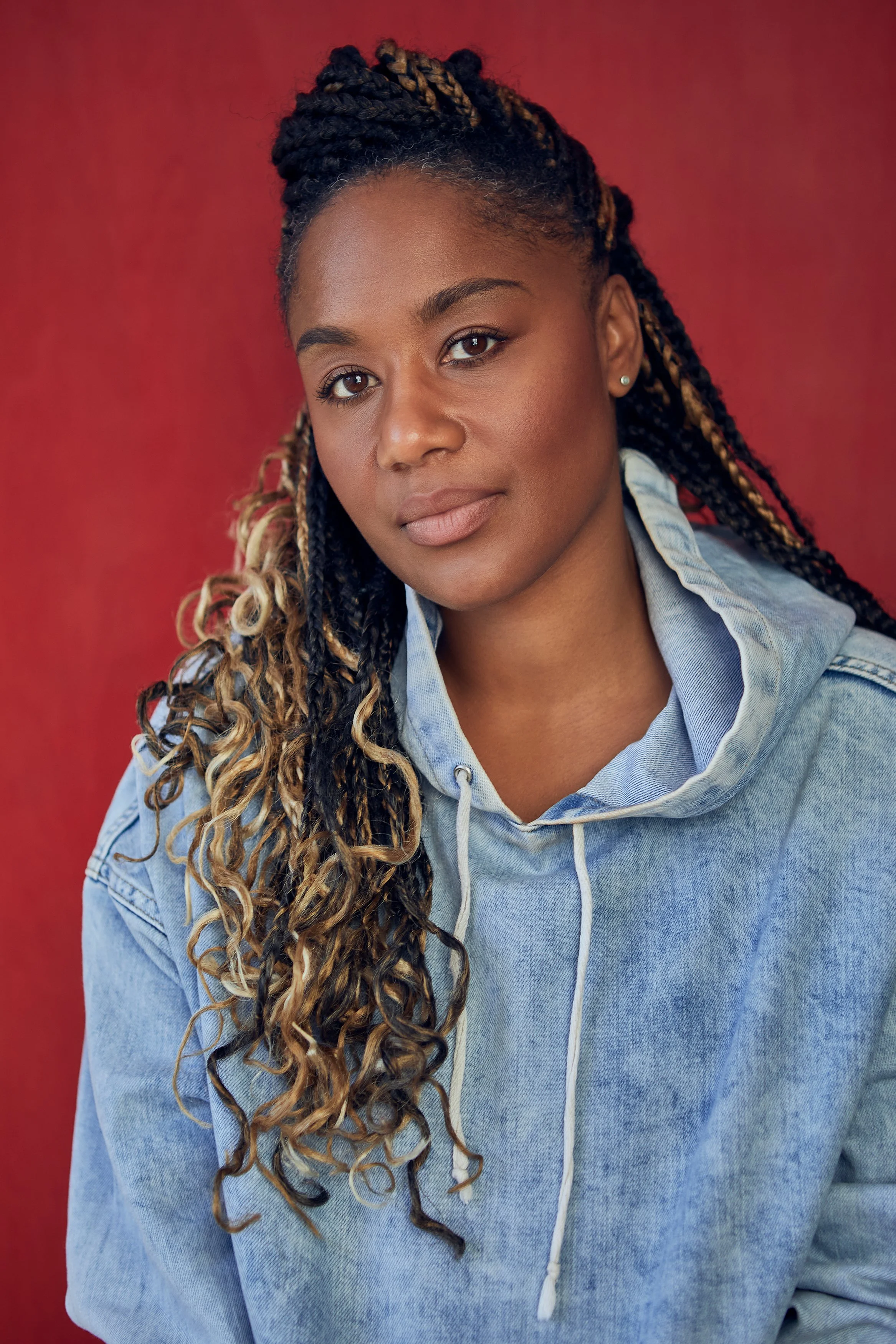 A young woman with dark skin and long braided hair with blonde highlights, wearing a light blue hoodie, looking at the camera, with a red background.