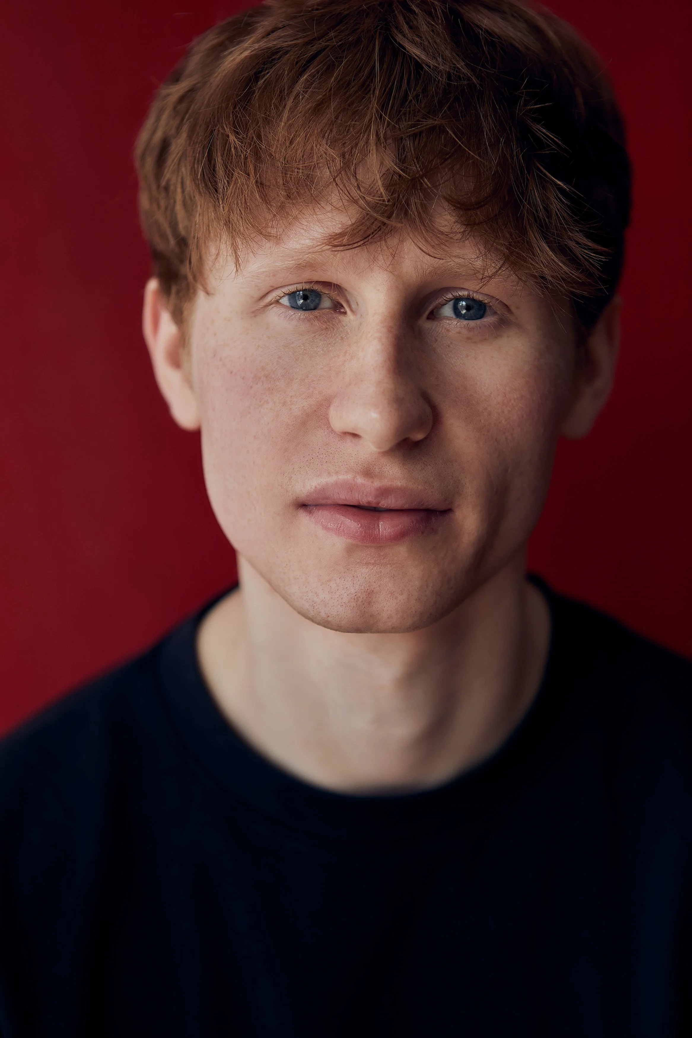 Close-up of a young man with red hair and blue eyes, wearing a dark shirt, against a red background.
