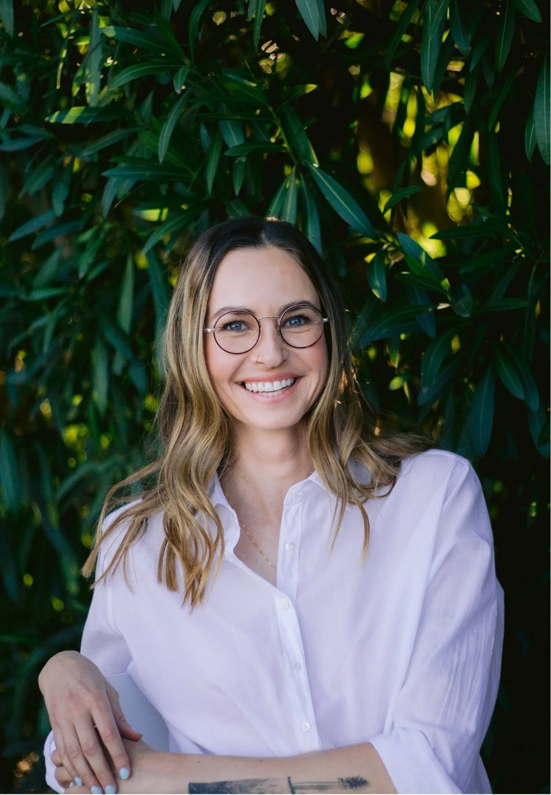 Adela Stone, a couples therapist, wearing glasses and a white button-up shirt, smiling outdoors in front of green foliage.