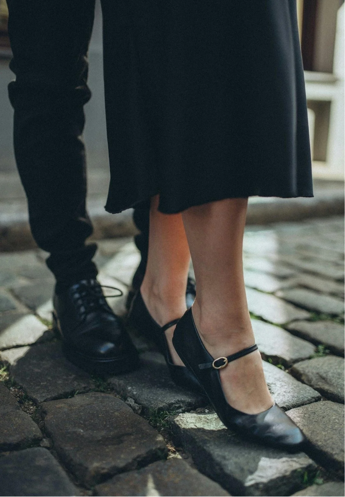Close-up of a woman's feet wearing black Mary Jane shoes, with a man standing behind her wearing black shoes and pants. They are on a cobblestone street. Representing a tight connection thanks to couples therapy