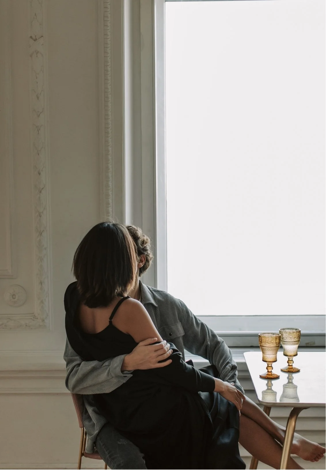 A woman sitting on a chair, next to a man, sharing an intimate moment in front of a large window, representing a renewed intimate connection following successful couples therapy sessions.