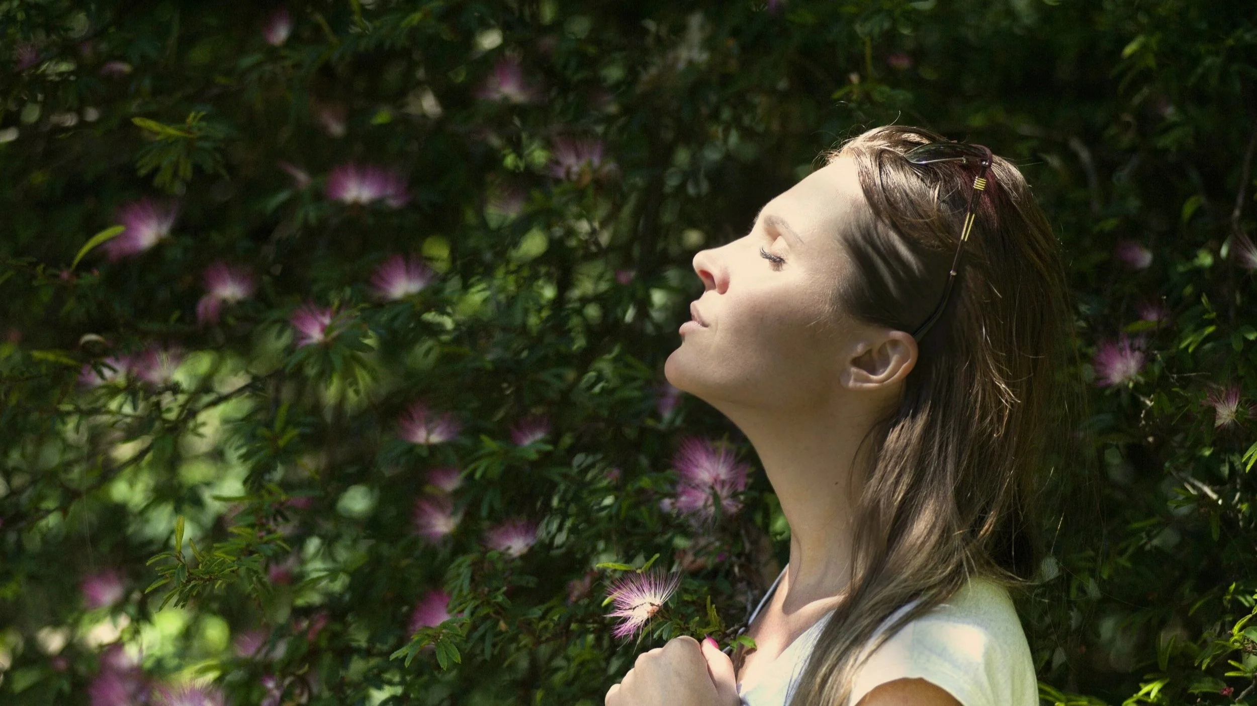 Woman with eyes closed, bathed in dappled light among trees, pausing to reconnect with herself and the natural world