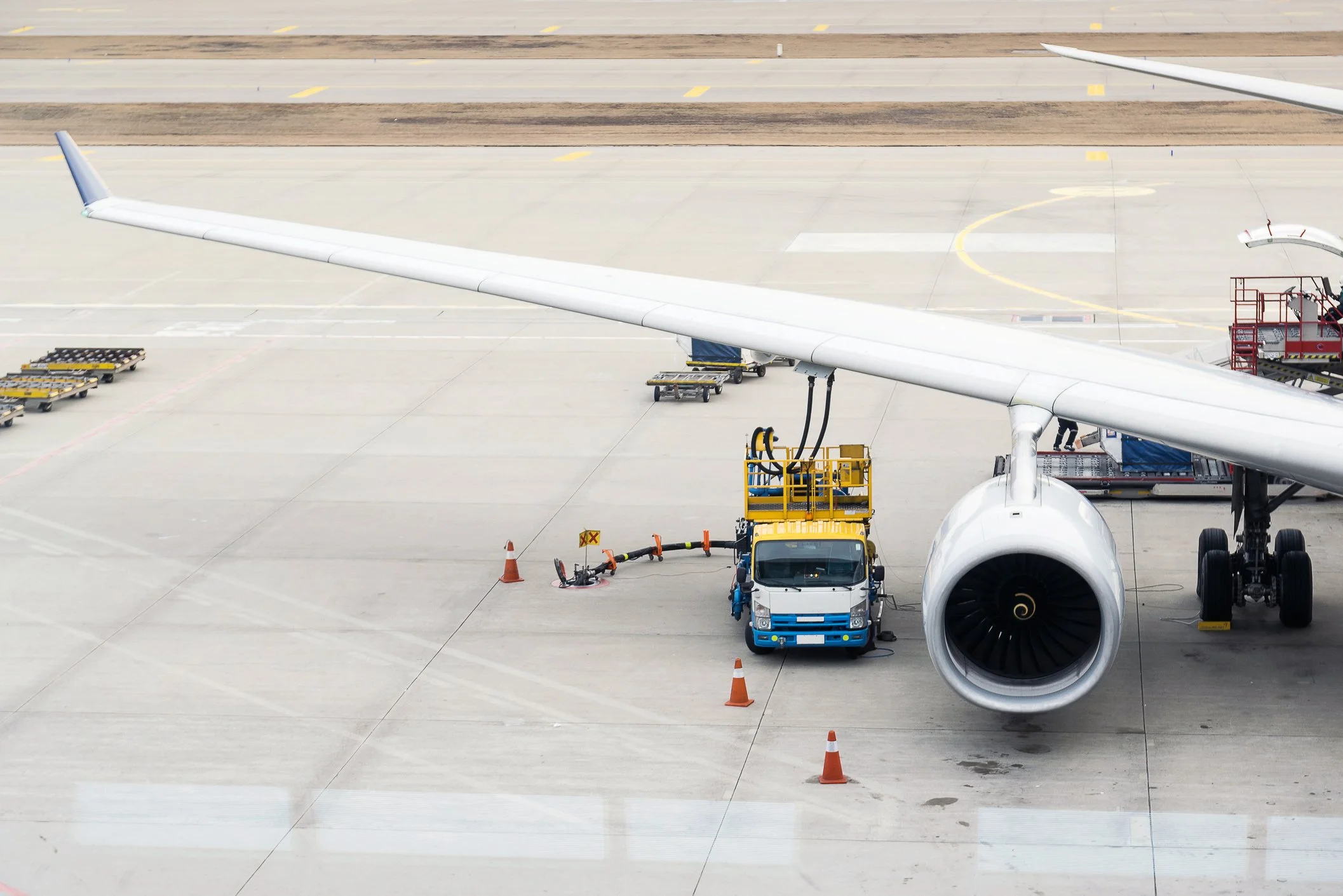Airplane at airport gate with jet engine visible, connected to ground service vehicle, orange cones around in a concrete apron.