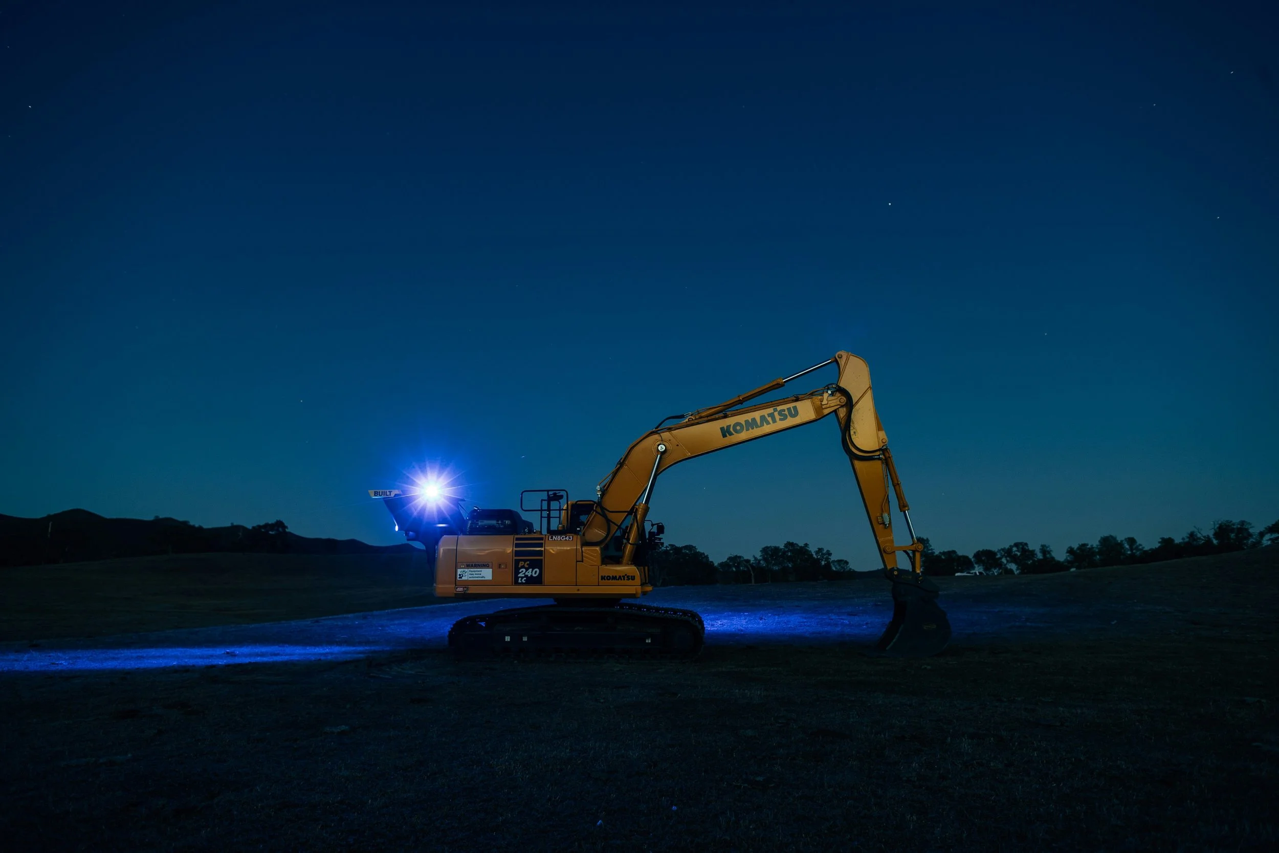 Yellow Komatsu excavator on a dark landscape at night with stars in the sky.