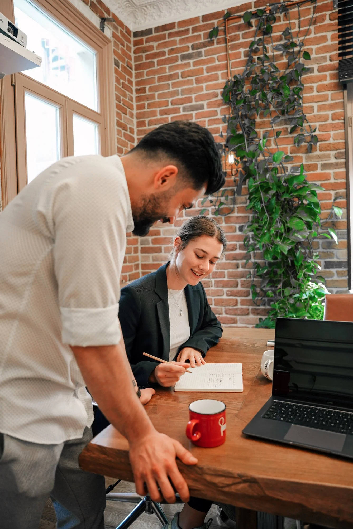 A man and a woman are having a discussion at a wooden conference table in a room with exposed brick wall and a large window. The woman is sitting and writing in a notebook, while the man is standing and leaning over the table, both smiling. There is a red mug, a laptop, and some office supplies on the table, and a large green plant is hanging on the brick wall.