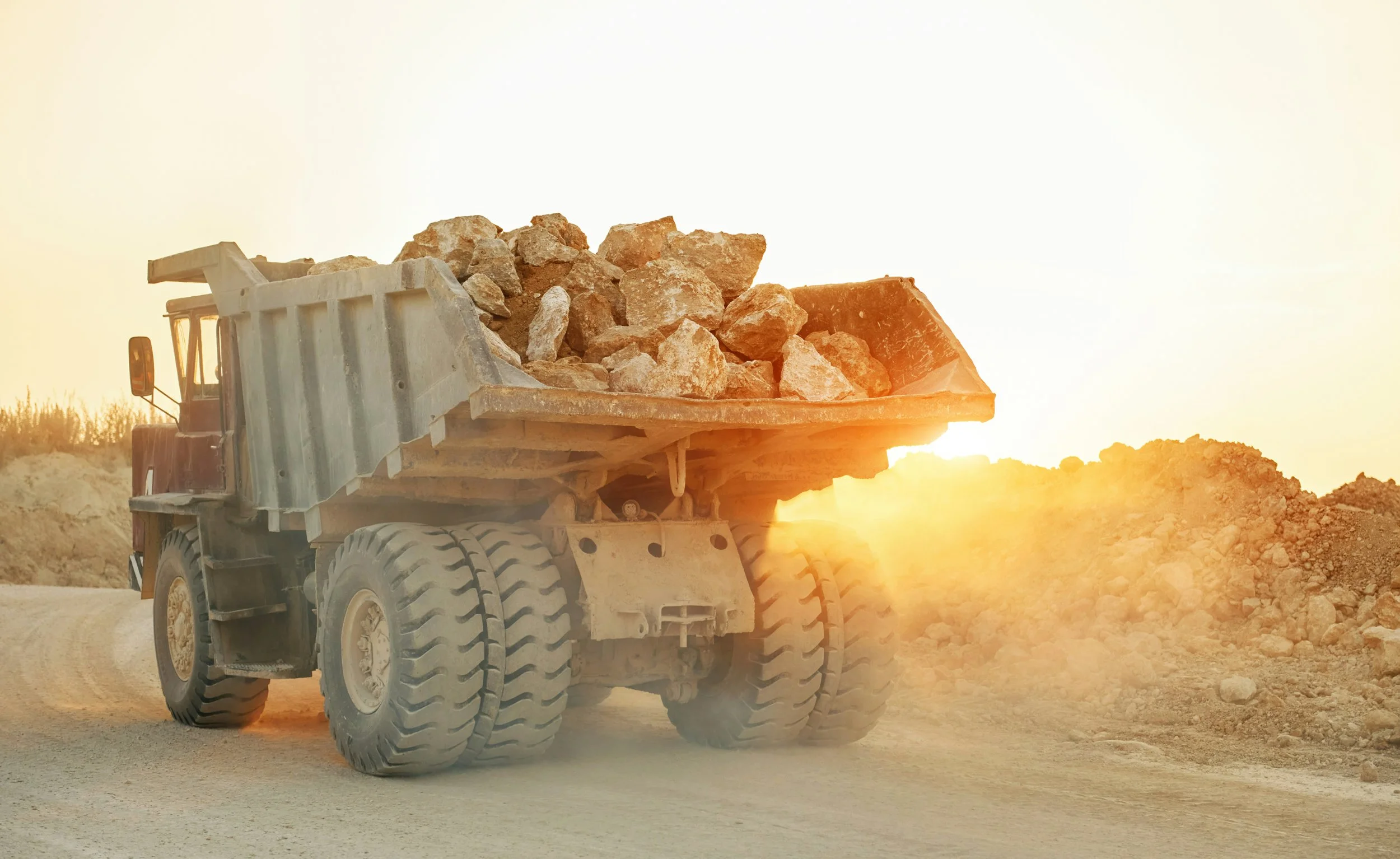 A large dump truck carrying rocks on a dirt construction site at sunset.