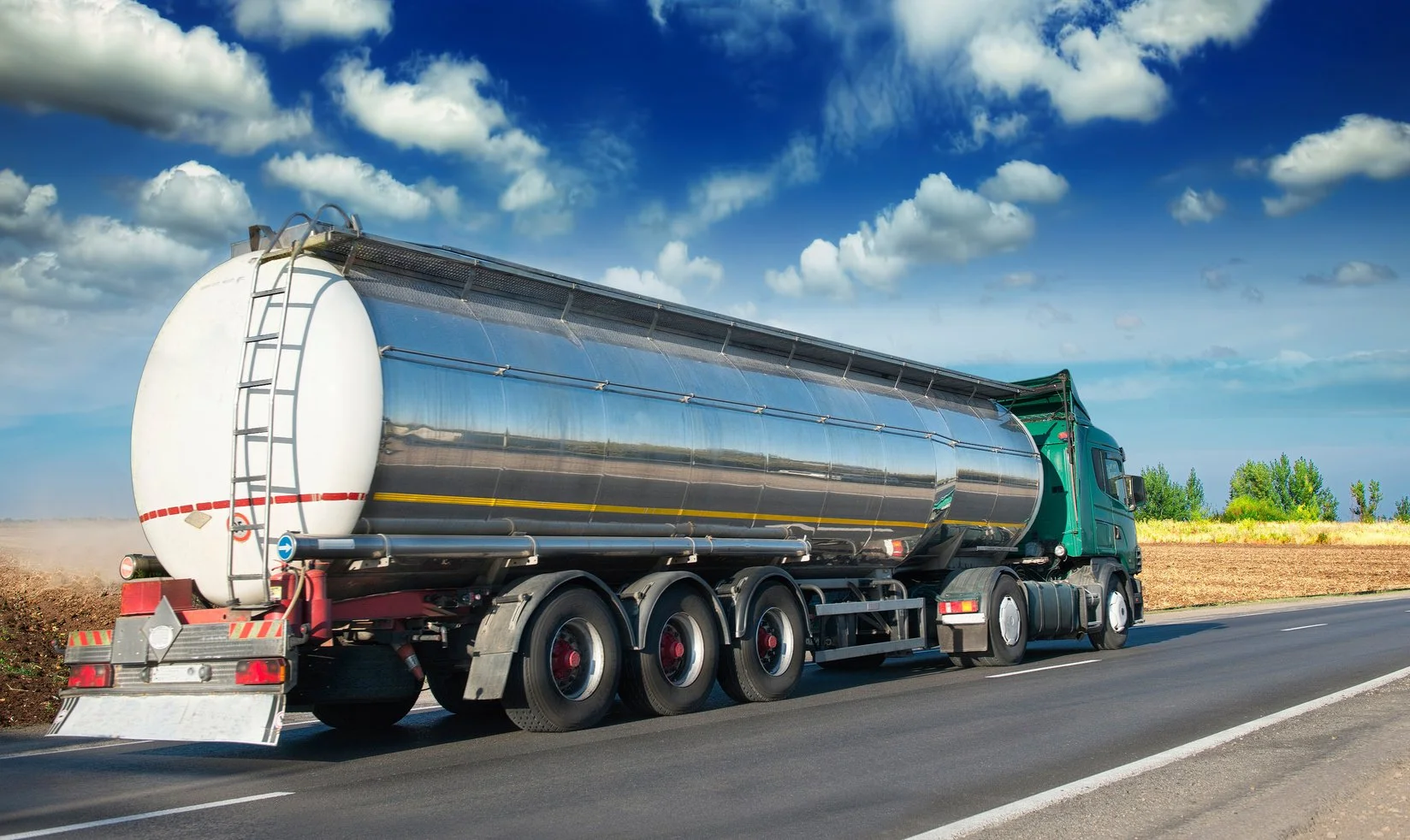 A green semi-truck with a shiny metallic tanker driving on a rural highway under a blue sky with scattered clouds.