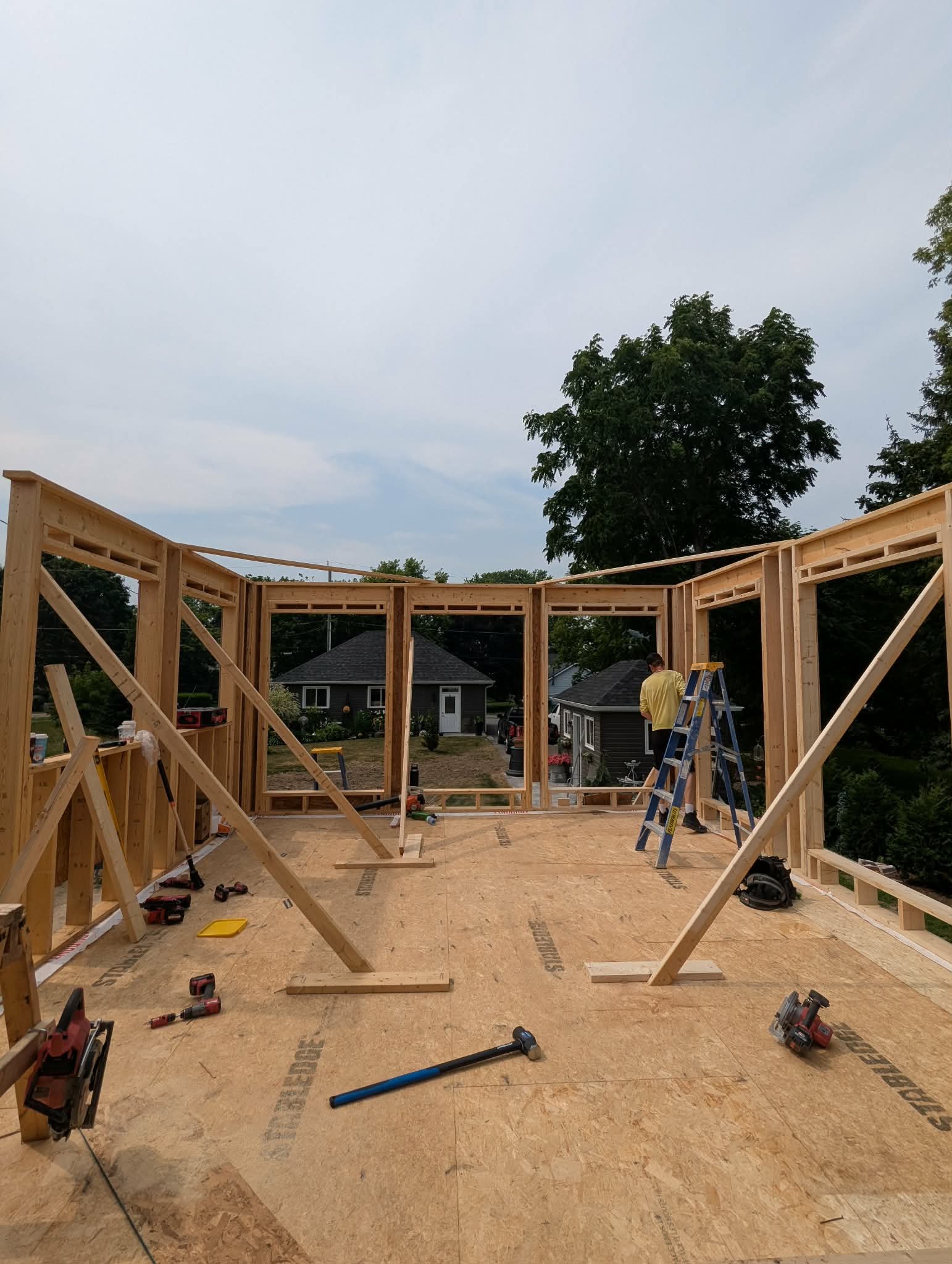 Construction site with wooden framing for a house, worker on ladder, tools, and nearby houses with trees.