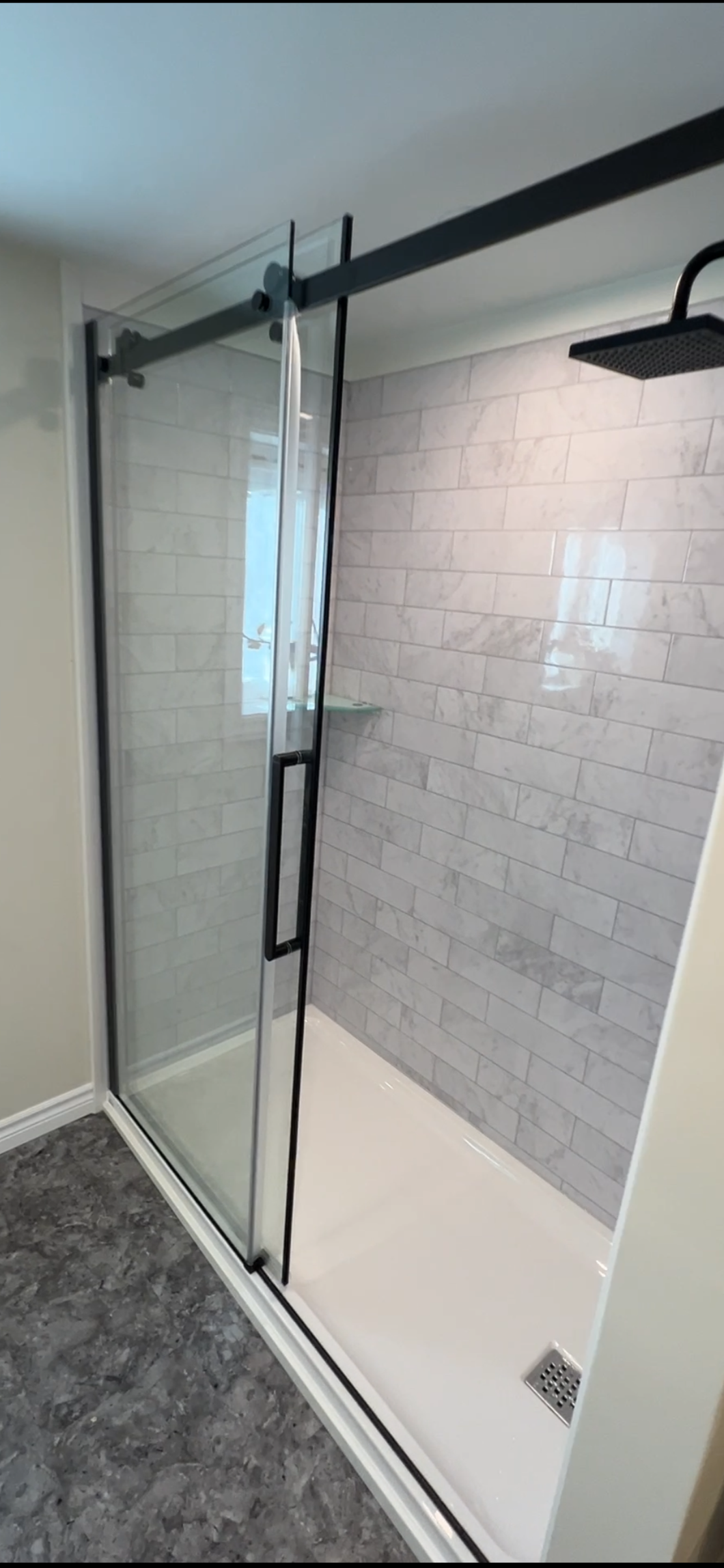 Modern shower with glass sliding door, grey subway tile walls, black showerhead, white base, and grey flooring.