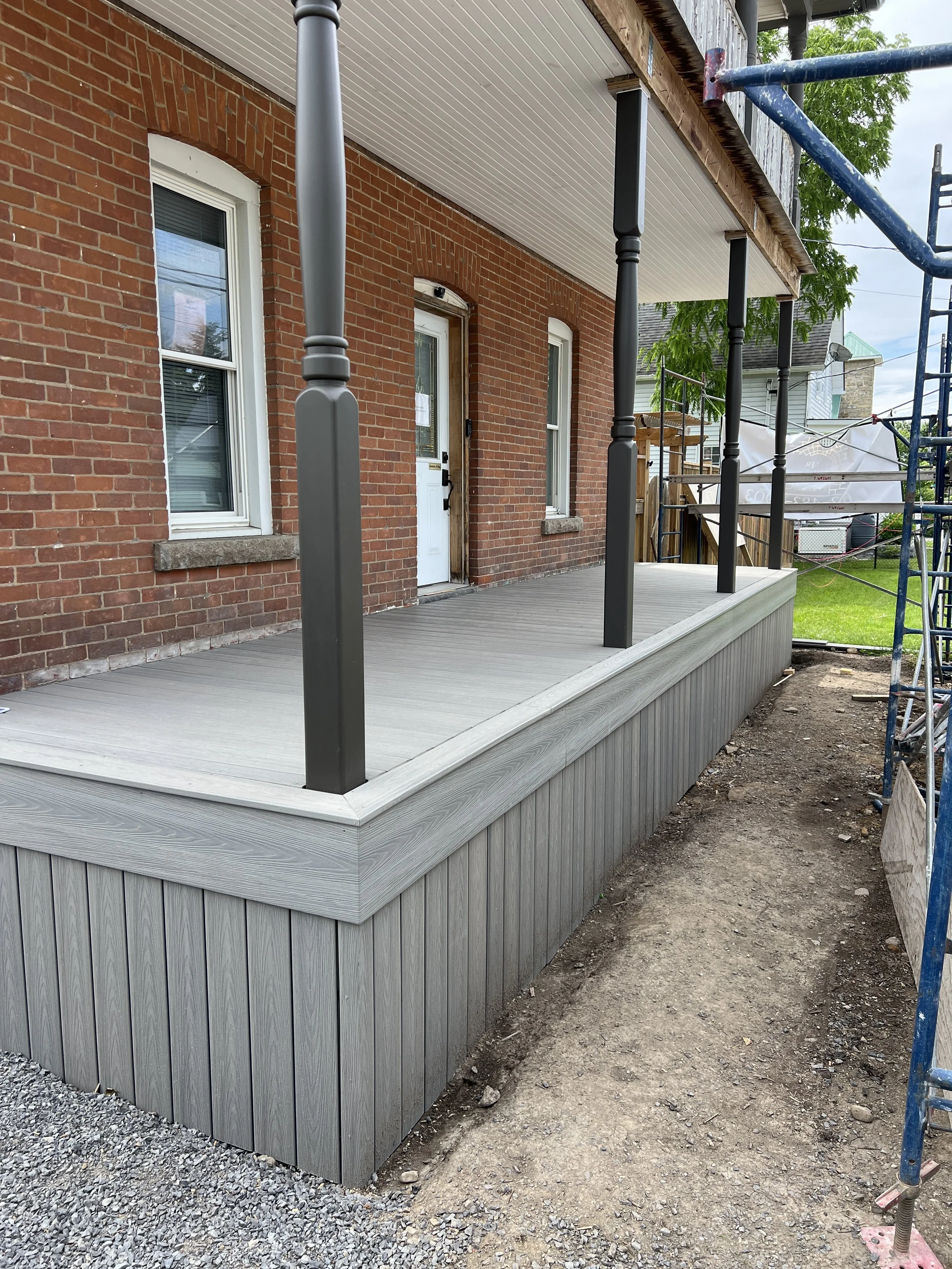 Construction of a new porch with gray decking and black metal railing supports attached to a brick house, with scaffolding on the right side.