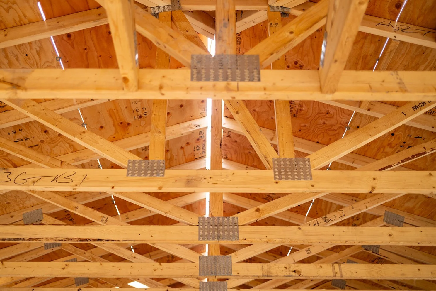 View of the ceiling of a building under construction, featuring wooden rafters and plywood sheathing, with visible metal plate connectors and small gaps allowing light to pass through.