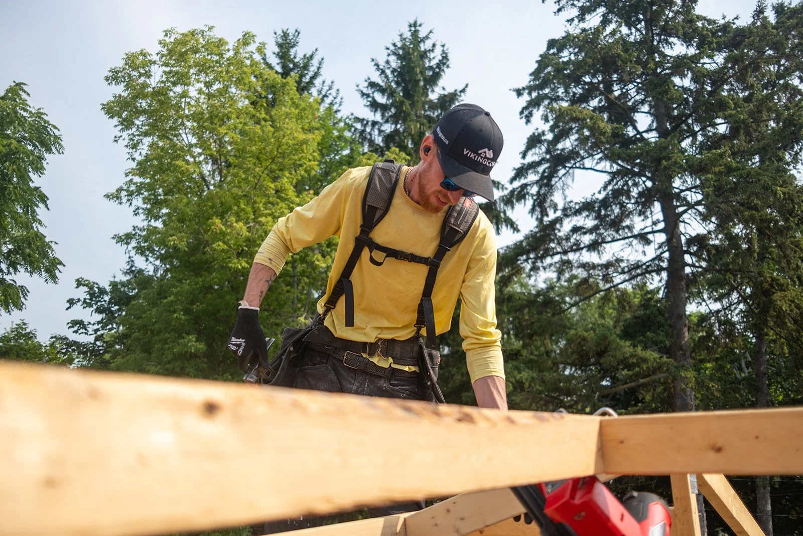 A man wearing a black cap, yellow long-sleeve shirt, black gloves, and sunglasses working on a wooden construction site outdoors with trees in the background.