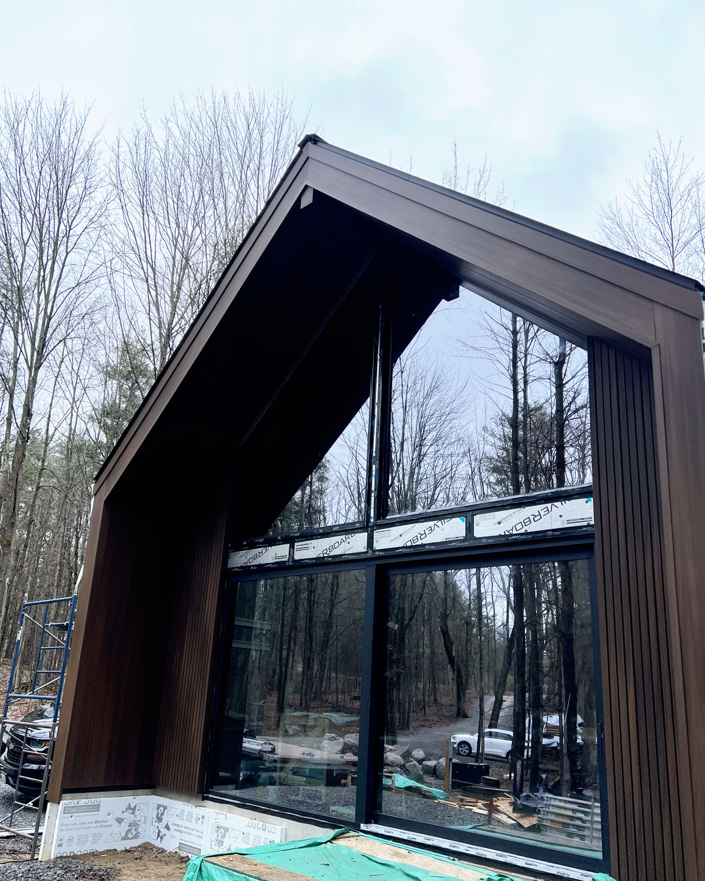 Construction site of a modern building with large glass windows and dark wood siding, surrounded by trees, with some construction materials and equipment visible.