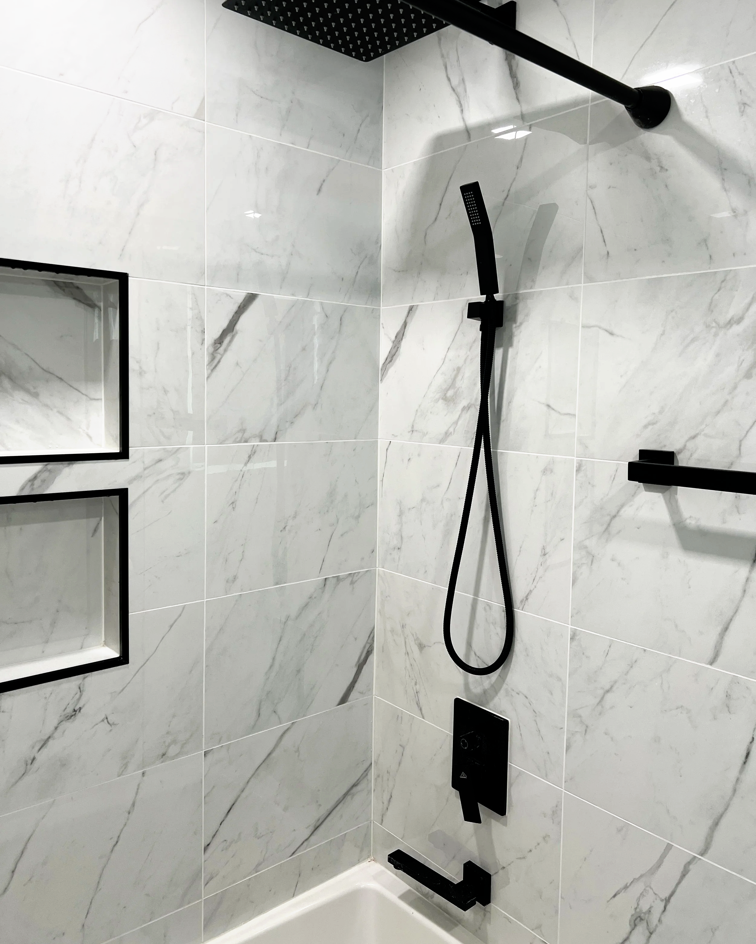 Modern shower with white marble tiles, black fixtures, a handheld showerhead, and built-in wall niches.