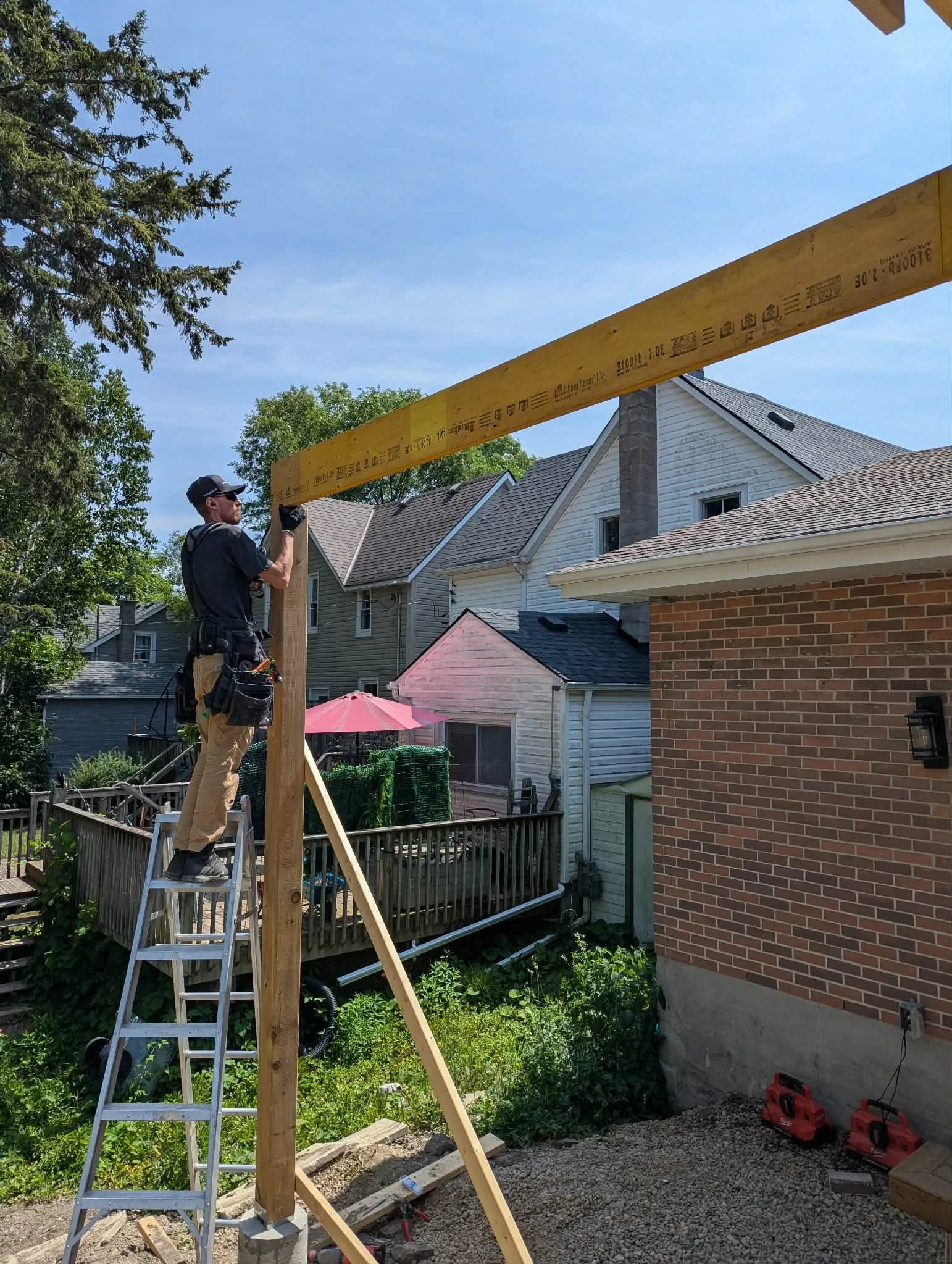 A man working on a construction project installing a horizontal wooden beam between vertical support posts outside a house, with a ladder, tools, and building materials visible.