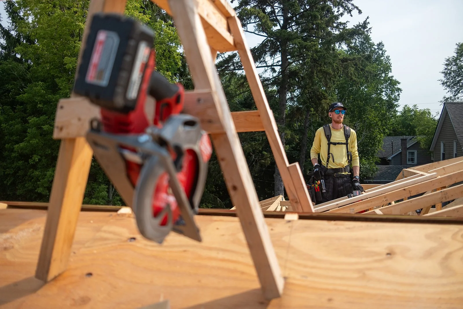 A construction worker stands on a wooden roof frame on a construction site, with a power saw on a wooden sawhorse in the foreground and trees and houses in the background.
