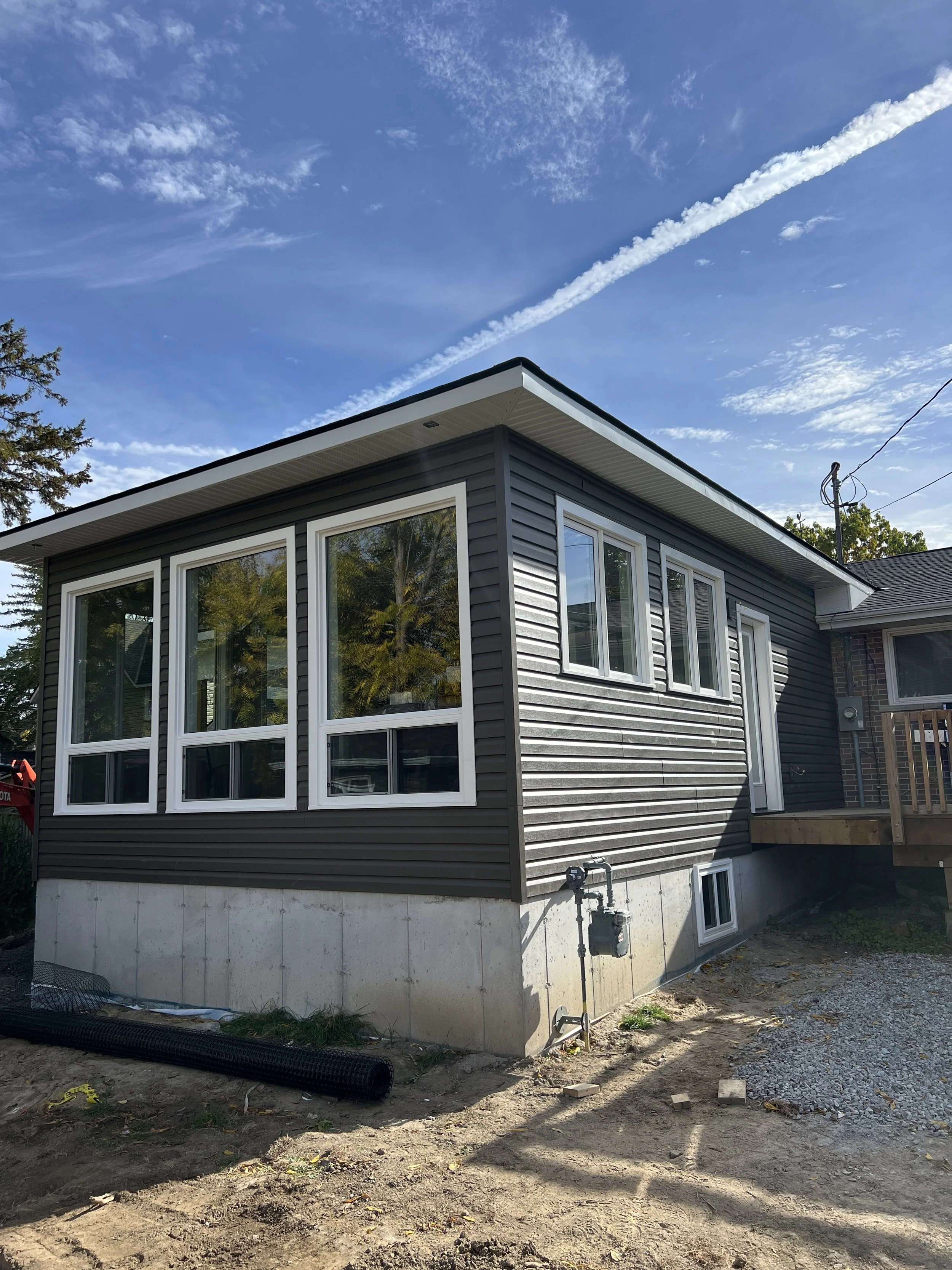 Newly constructed house with black siding, large front windows, and a small side window, elevated on a concrete foundation. There is a wooden deck attached on one side, and the ground around is unfinished with dirt and gravel. Overhead, a blue sky wi