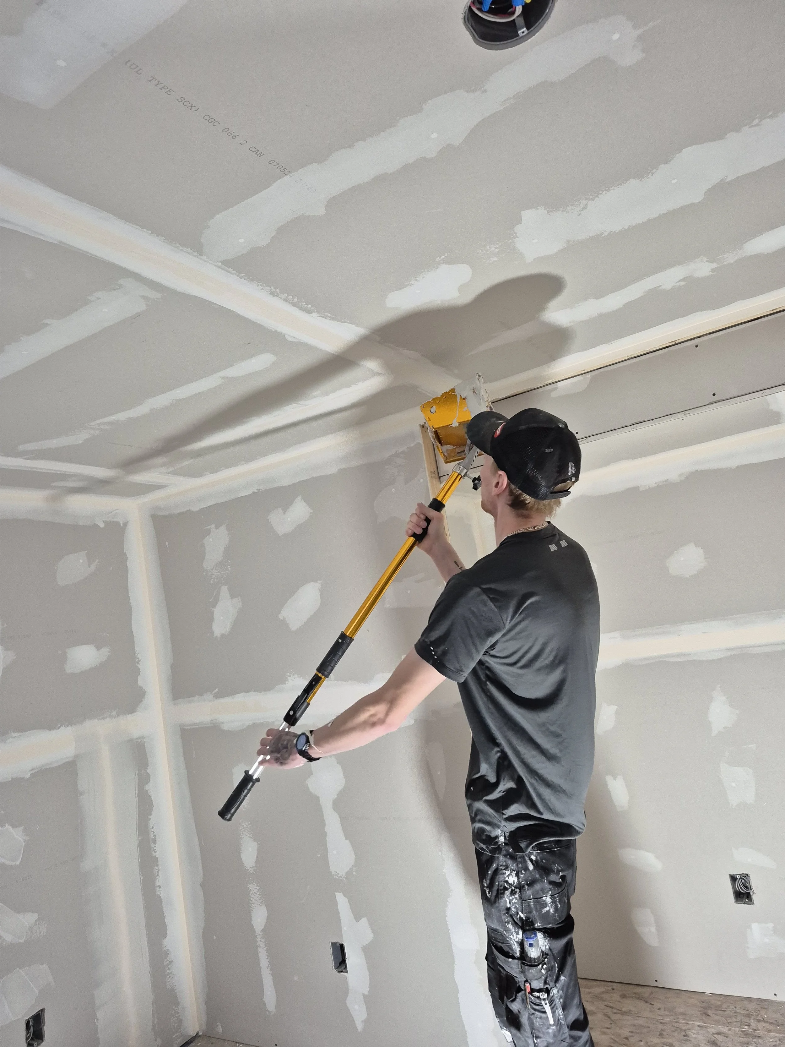A man wearing a black cap, black t-shirt, and paint-splattered pants is using a drywall sander on a finished drywall ceiling in a room under construction.