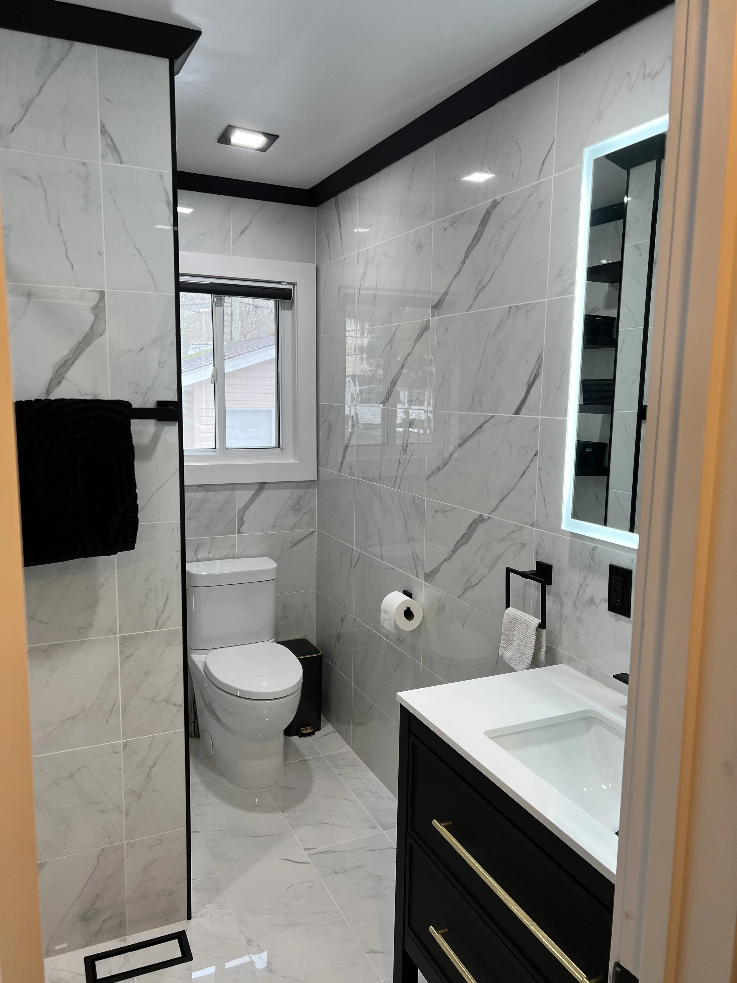 Modern bathroom with white marble walls and floor, a toilet, a black vanity with gold handles, a mirror with LED lighting, and a small window.