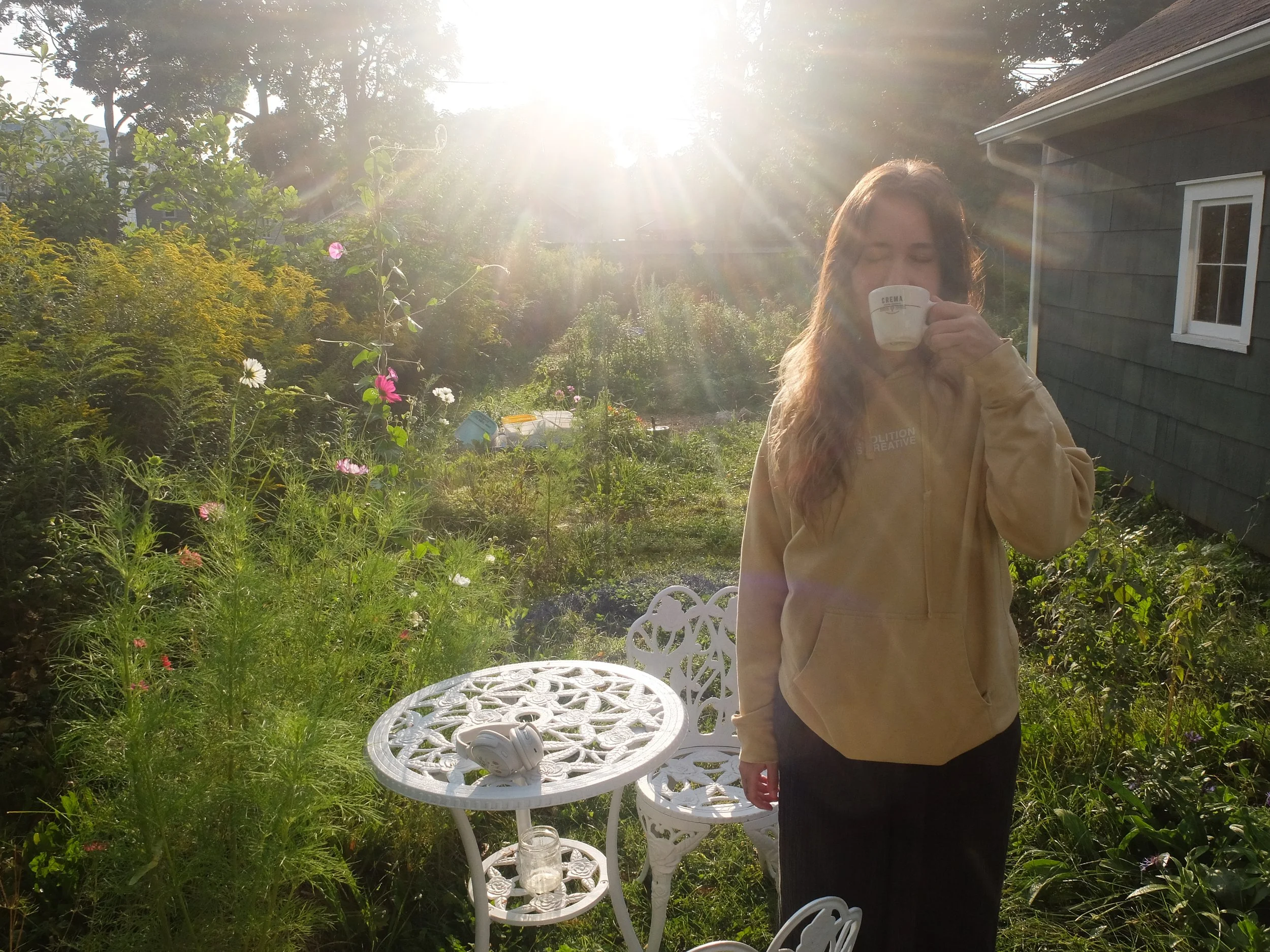 woman drinking coffee facing camera very sunny garden. a woman with a long brown hair. woman in garden sunny orange hoodie coffee.