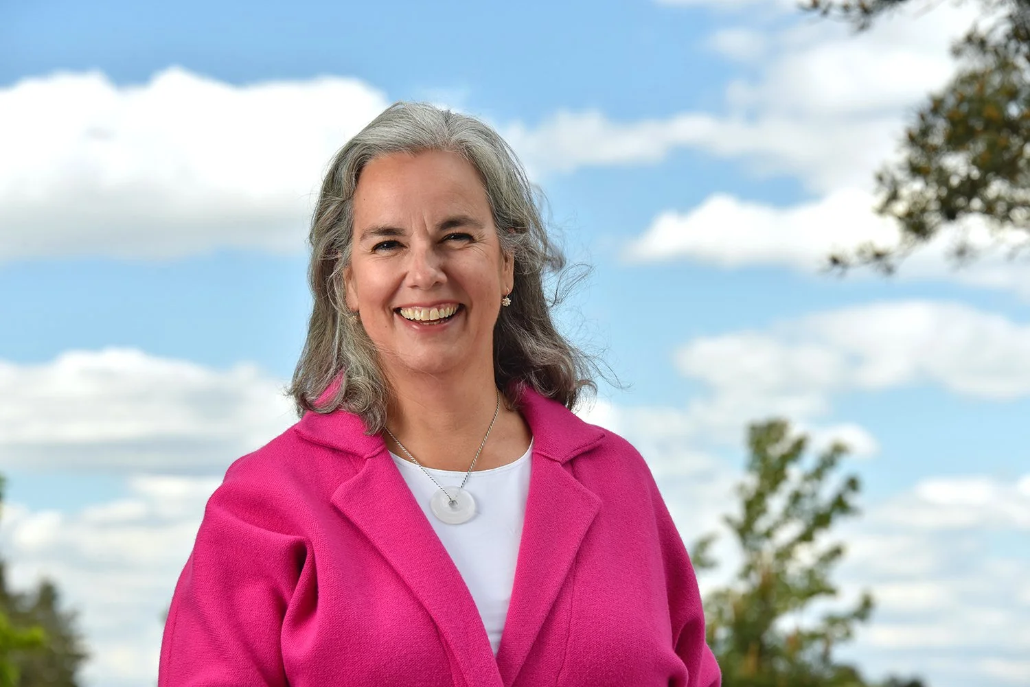 Marike smiling outdoors, wearing a pink coat and a white top, with trees and a cloudy sky in the background.