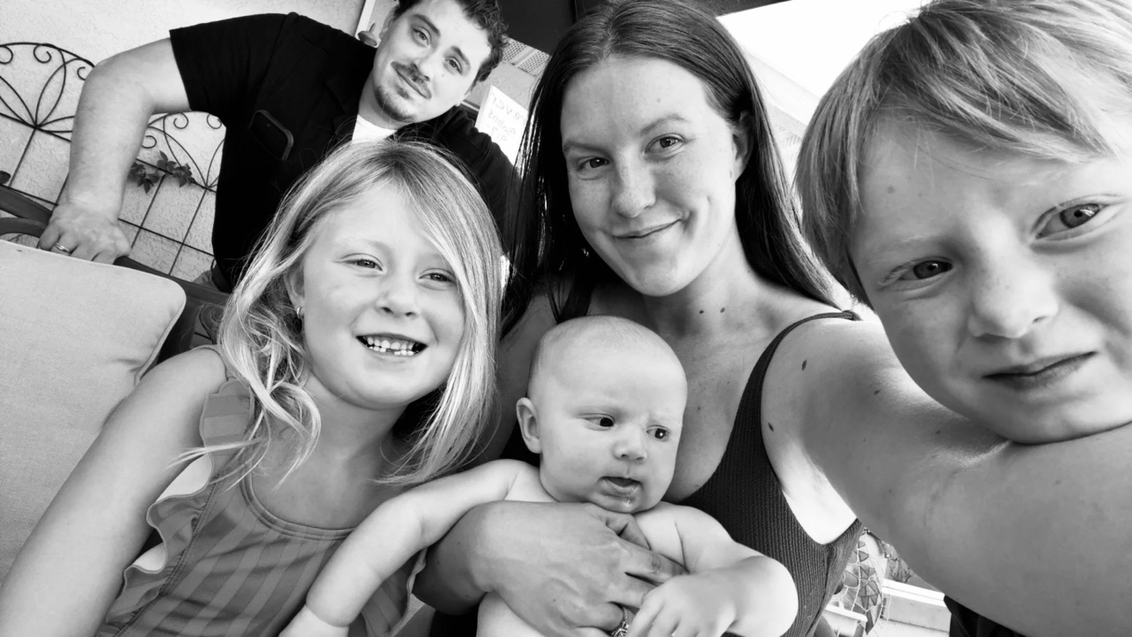A black-and-white photo of a family taking a selfie. There are five people: a smiling young girl with long hair, a baby being held by a woman with straight hair, a child with a slight smile and freckles, a wide-mouthed man in the background leaning over a table, and a young boy with a big smile on the right side of the photo.
