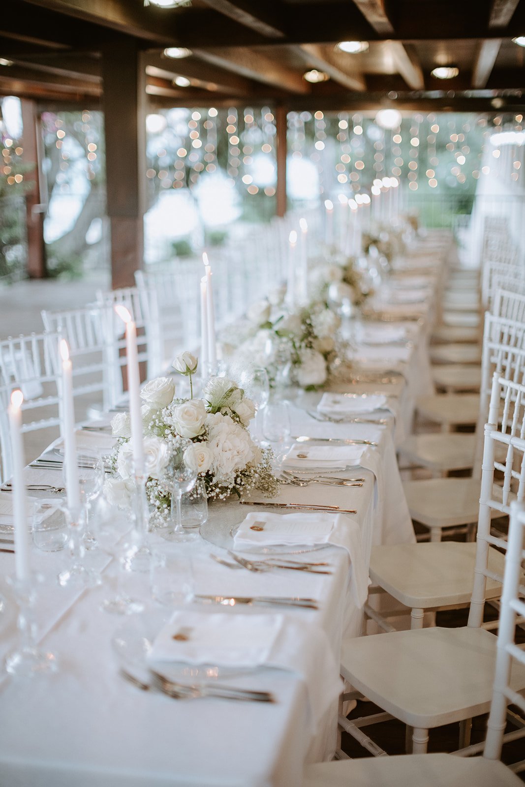 Elegant wedding reception table decorated with white flowers, candles, and place settings, under string lights in a wooden pavilion.