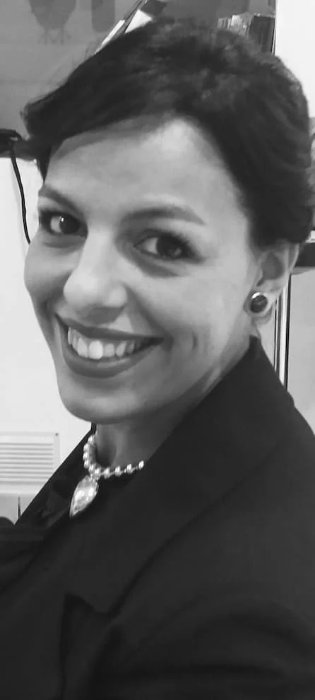 Black and white photo of a woman smiling, wearing earrings, a pearl necklace with a large pendant, and a jacket.