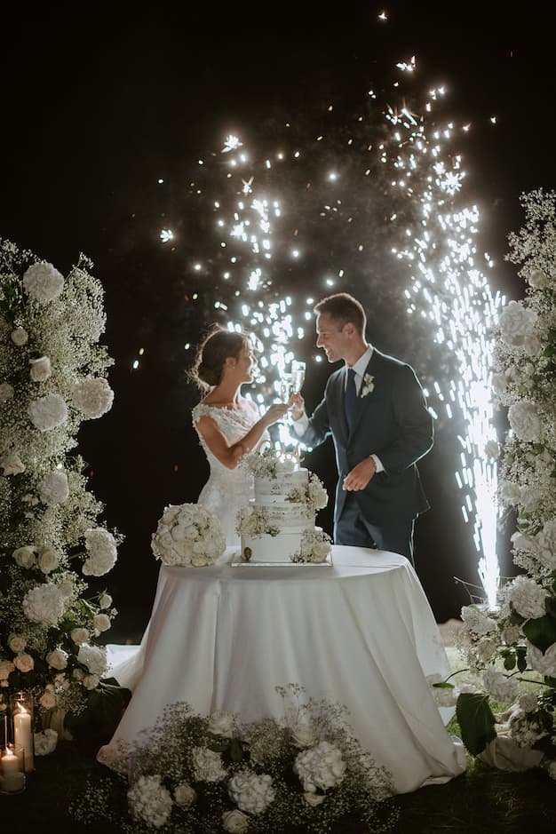 A bride and groom celebrate with a toast at their wedding reception, surrounded by white floral arrangements and fireworks display in the background.