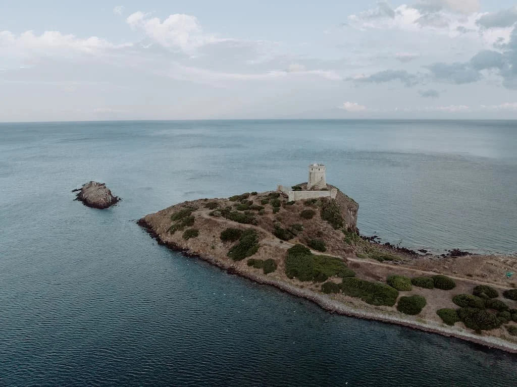 Aerial view of a coastal area with a small island featuring a tower on a hill and another small rock formation nearby in the ocean.