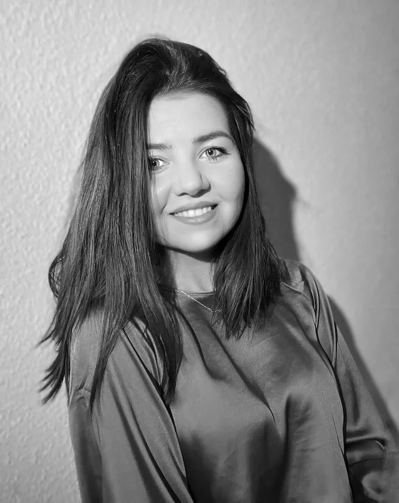 A black and white portrait of a young woman with shoulder-length hair, smiling and wearing a shiny top, standing against a wall with a textured surface.