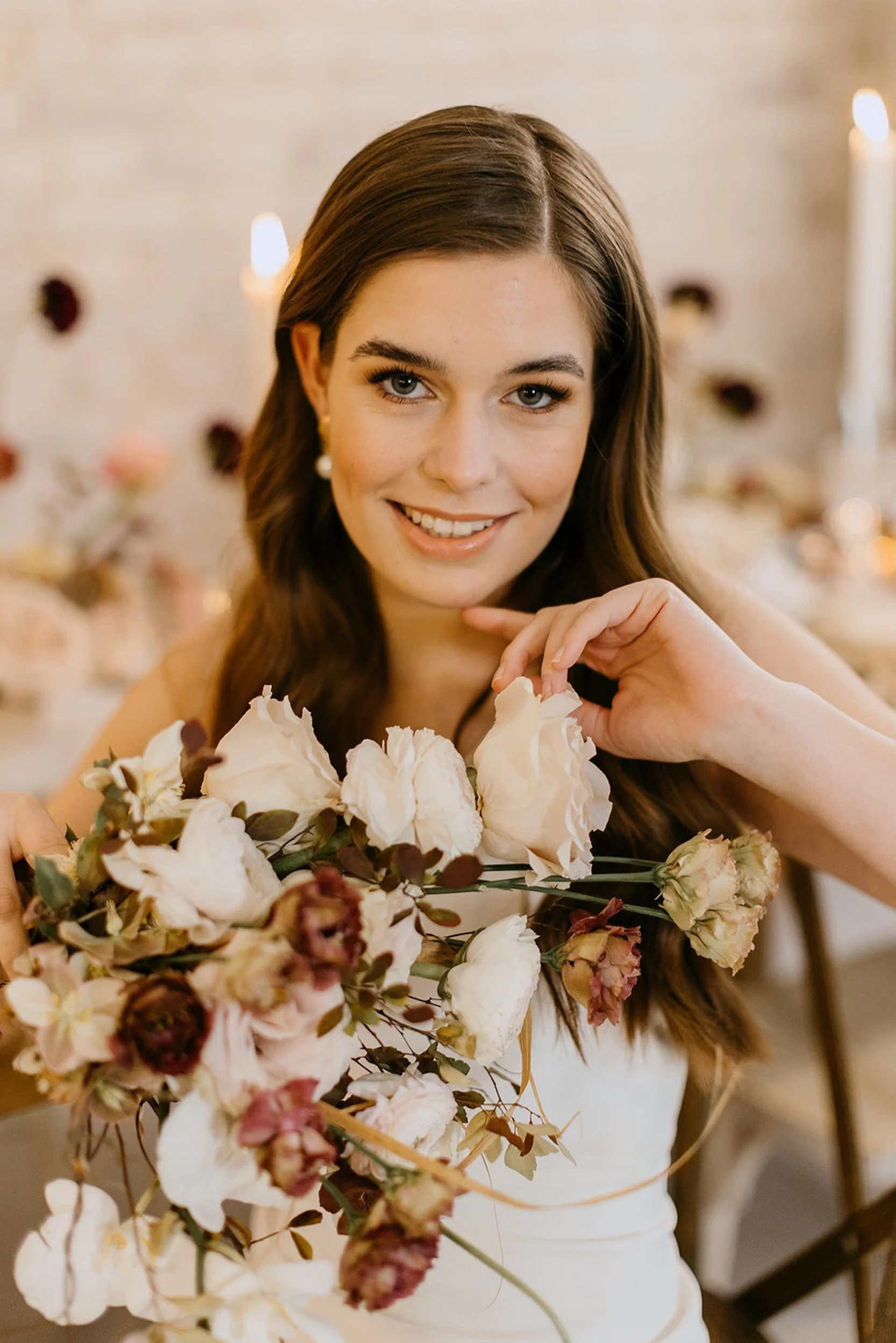 A young woman with long brown hair in a wedding dress holds a bouquet of white and pink flowers, smiling at the camera.
