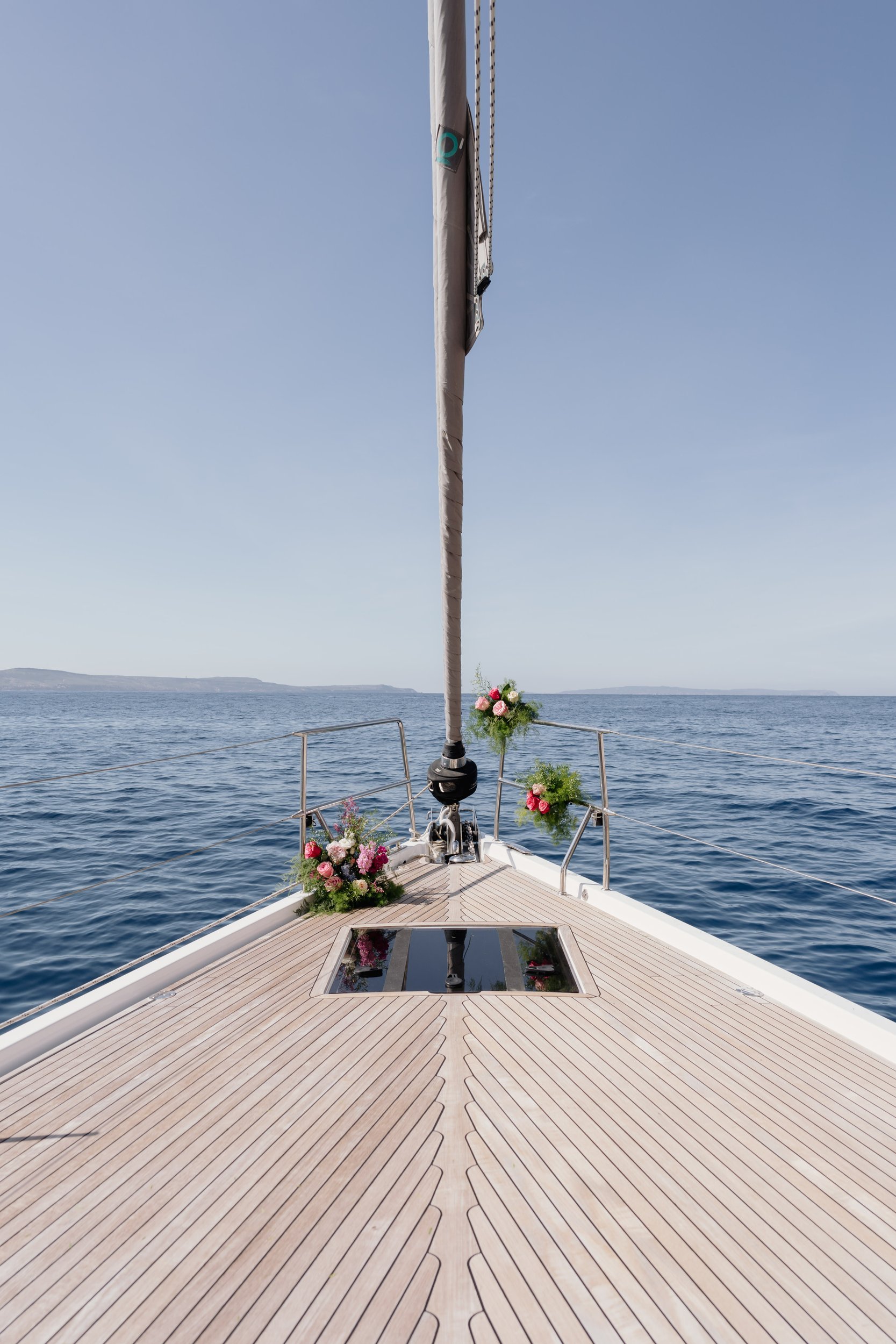 View from the bow of a sailboat decorated with pink and white flowers, sailing on calm blue waters of Sardinia under clear sky, with islands in the distance.