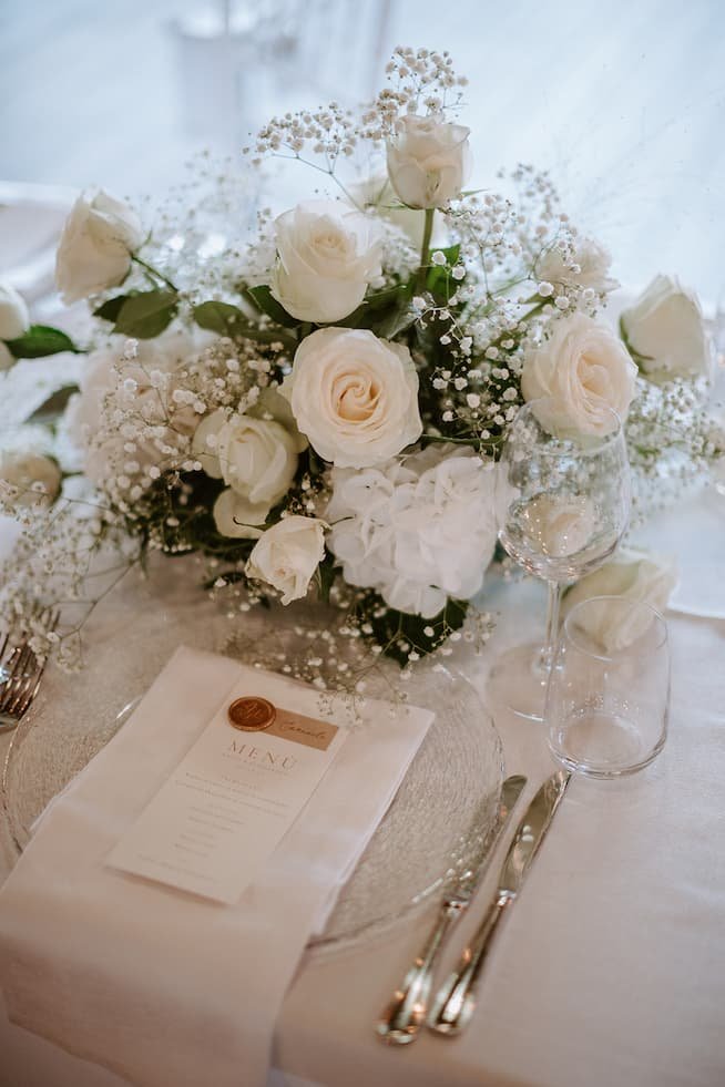 Elegant table setting featuring a floral centerpiece with white roses and baby's breath, wine glasses, a menu with a wax seal, and silverware on a white tablecloth.
