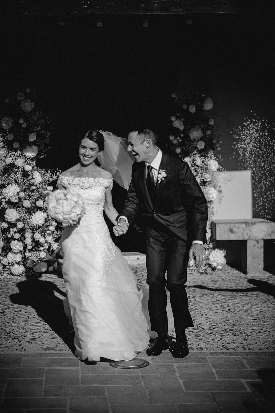 A bride and groom smiling and holding hands, celebrating their wedding indoors with floral decorations in the background, in a black and white photo.
