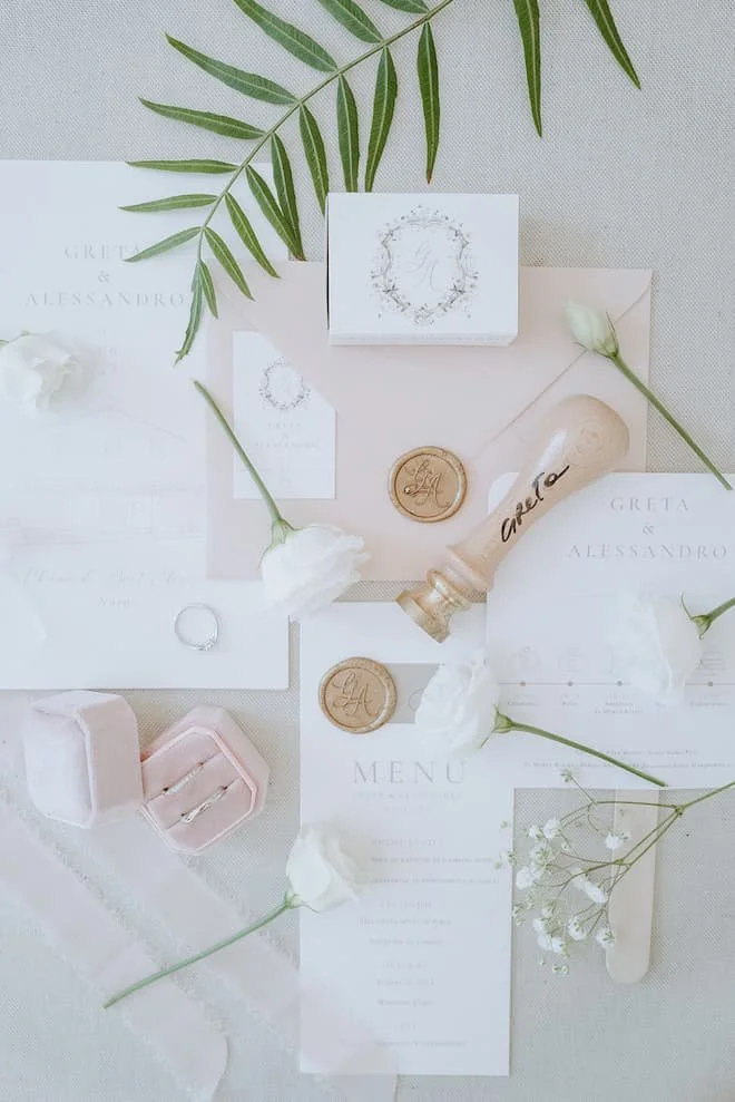 Wedding invitation suite with floral accents, wax seals, and a wax stamp, surrounded by white flowers and greenery in soft pastel tones.