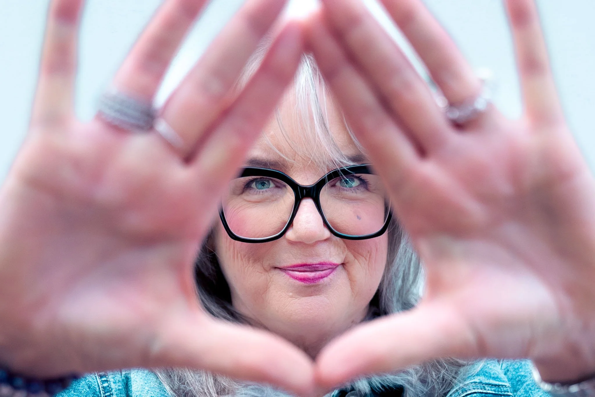 A woman with glasses and gray hair looking through her hands, which are framing her face, with a light blue background.