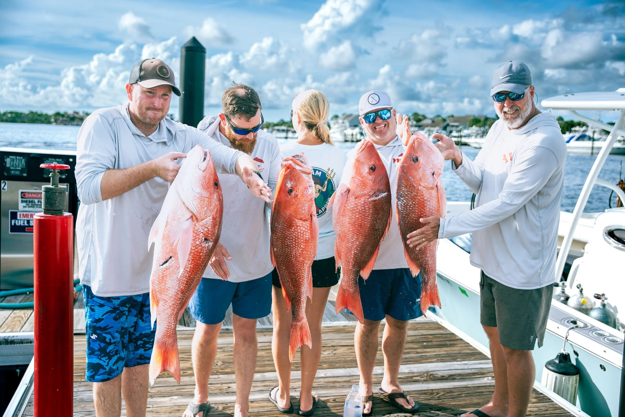 Group of five people on a boat dock at Point South Marina - Bay Point (Panama City Beach) holding up red snapper fish after a fishing trip, with boats and a cloudy sky in the background.