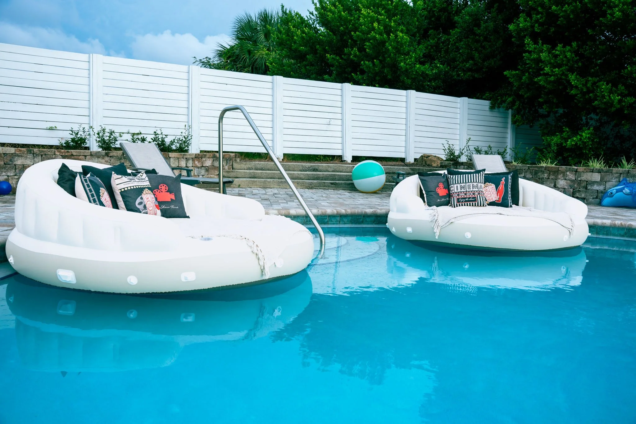 A swimming pool with two inflatable white pool floats, each topped with decorative pillows, a ball in the water, a white fence, green trees, and a cloudy sky in the background.