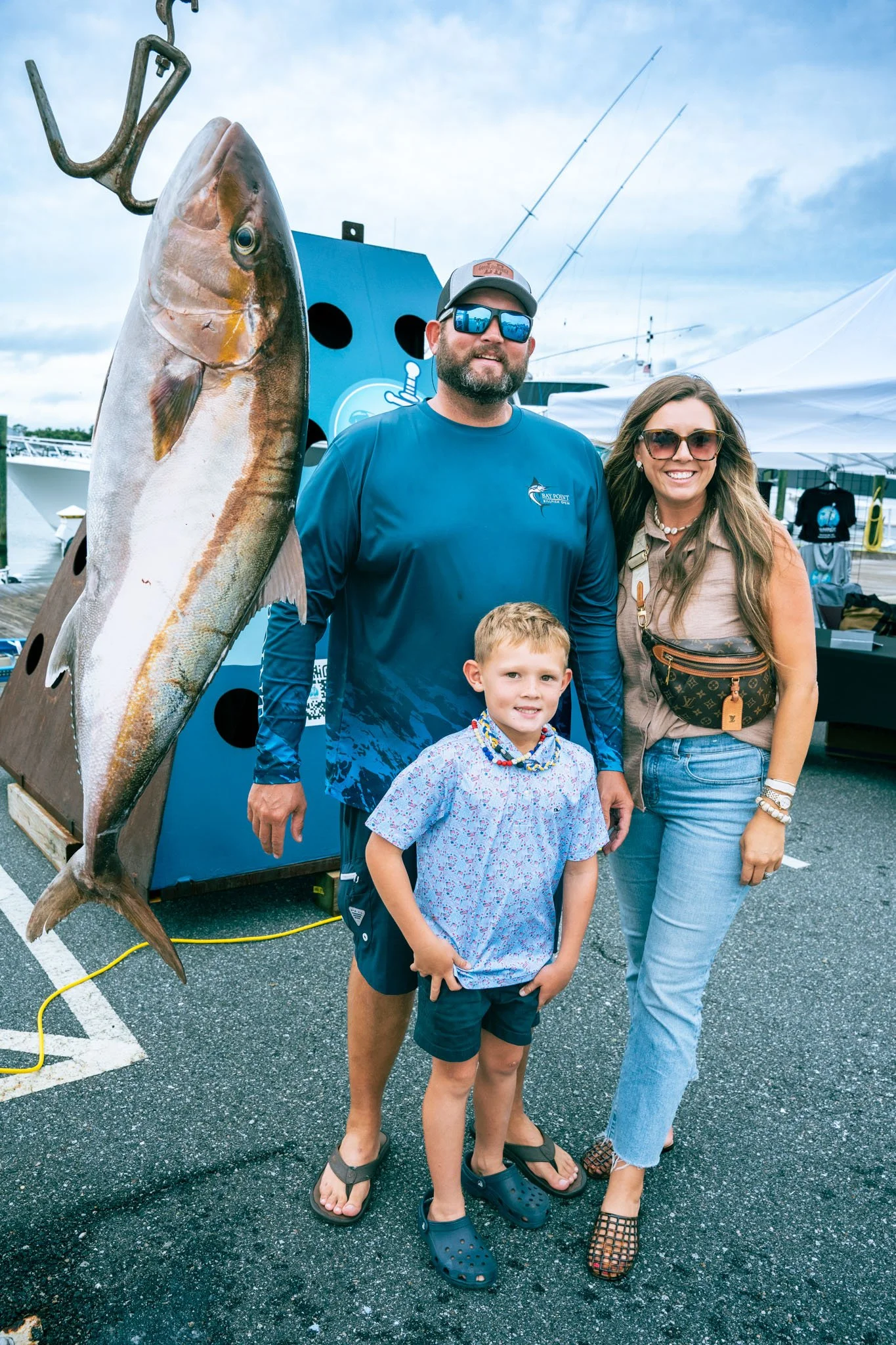 A man and woman stand with two children at a dock, posing with a large fish on a fish display stand. The man is wearing sunglasses, a baseball cap, and a blue long-sleeve shirt. The woman is wearing sunglasses, a beige top, and jeans. One child is we