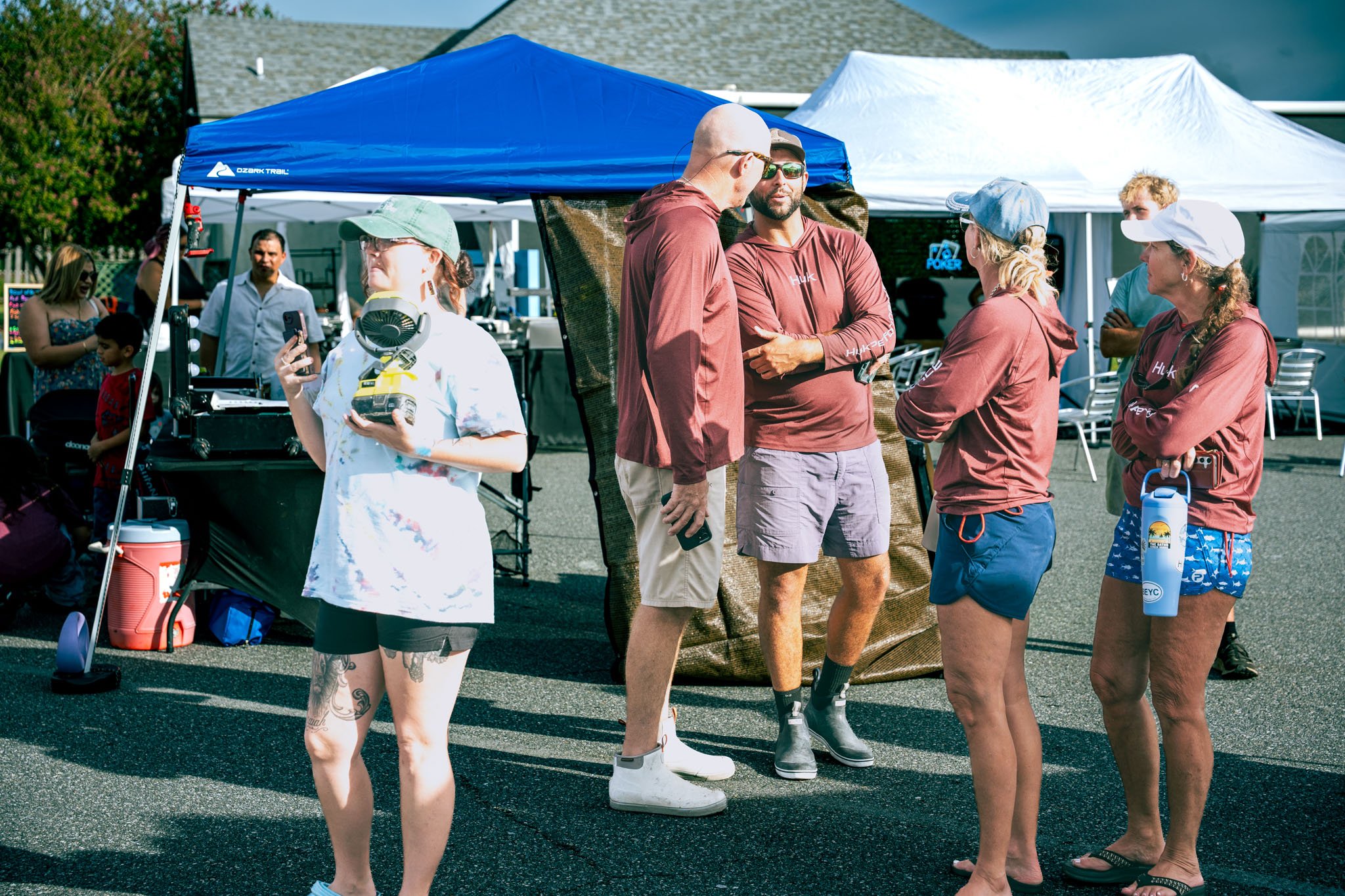 Group of people standing and talking outdoors near tents at the Warrior Memorial Reefs Reels and Spears Fishing Tournament event at Point South Marina - Bay Point (Panama City Beach), with some holding items like water bottles and cell phones.