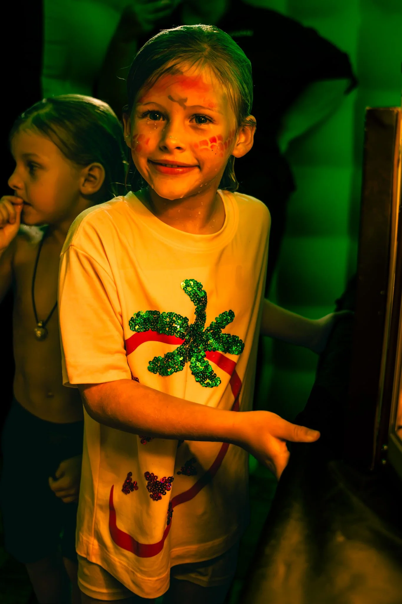 Smiling young girl with face paint, wearing a yellow tee with a glittery green dragonfly design, in a party or event setting with people and green lighting.