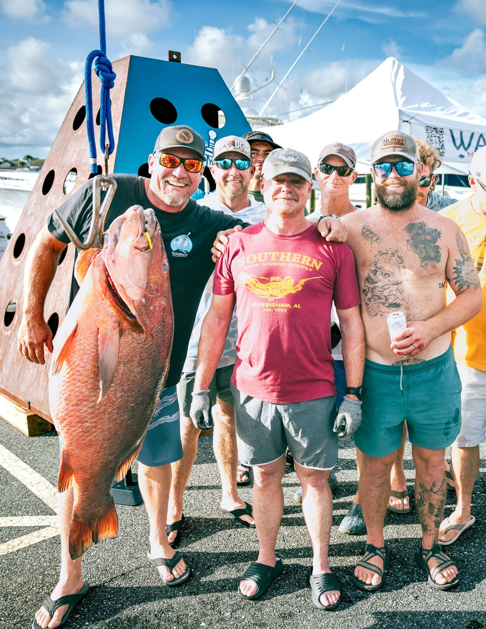 Group of men standing together at the Warrior Memorial Reefs Reels and Spears Fishing Tournament event at Point South Marina - Bay Point (Panama City Beach), with one holding a large cubera snapper and a wooden structure behind them.