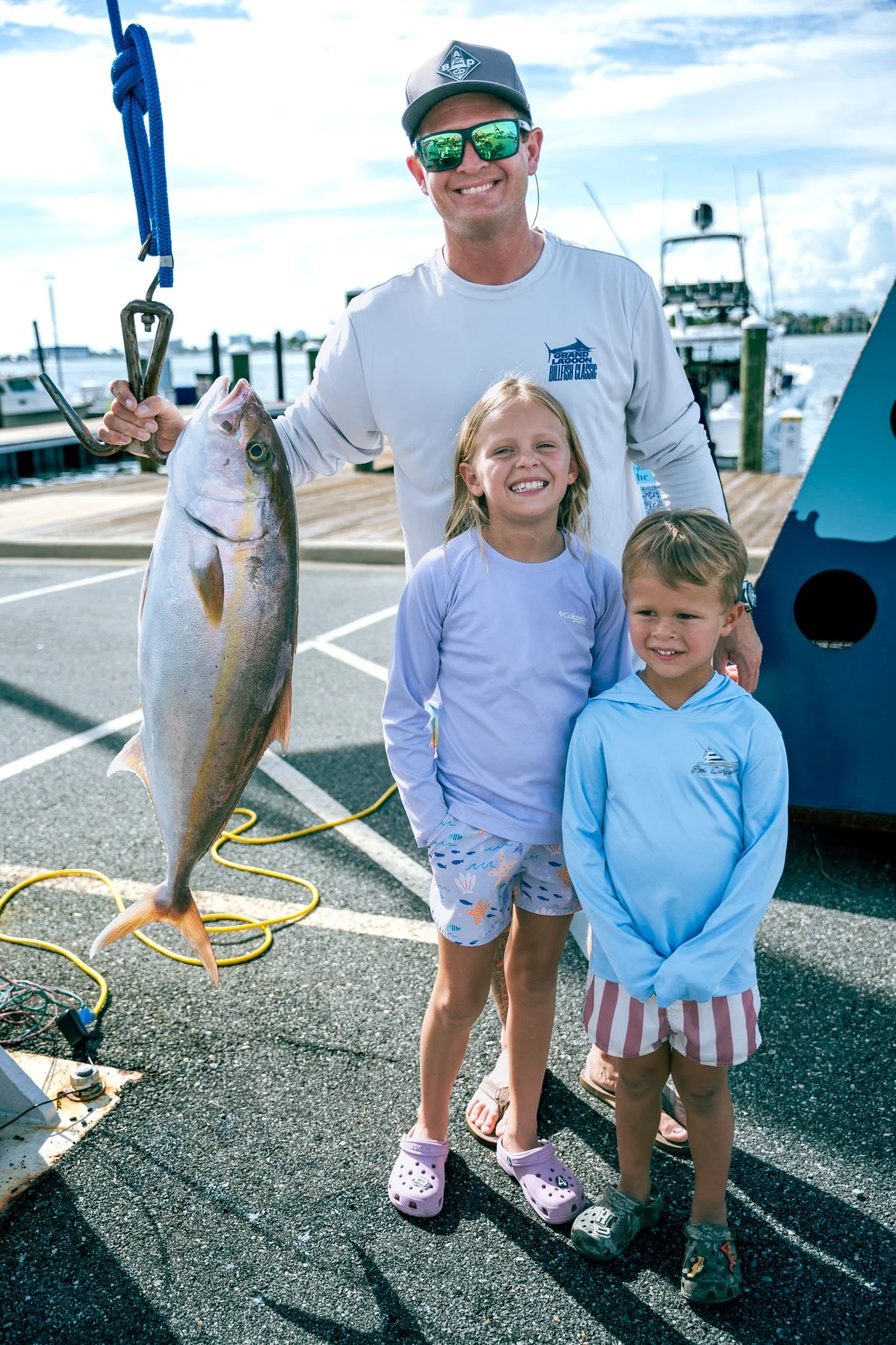 A smiling man wearing sunglasses and a baseball cap holds a large fish with a fish gripper, standing on a dock with two children in front of him. The children, a girl and a boy, are smiling and wearing casual clothing. Several boats are visible in th