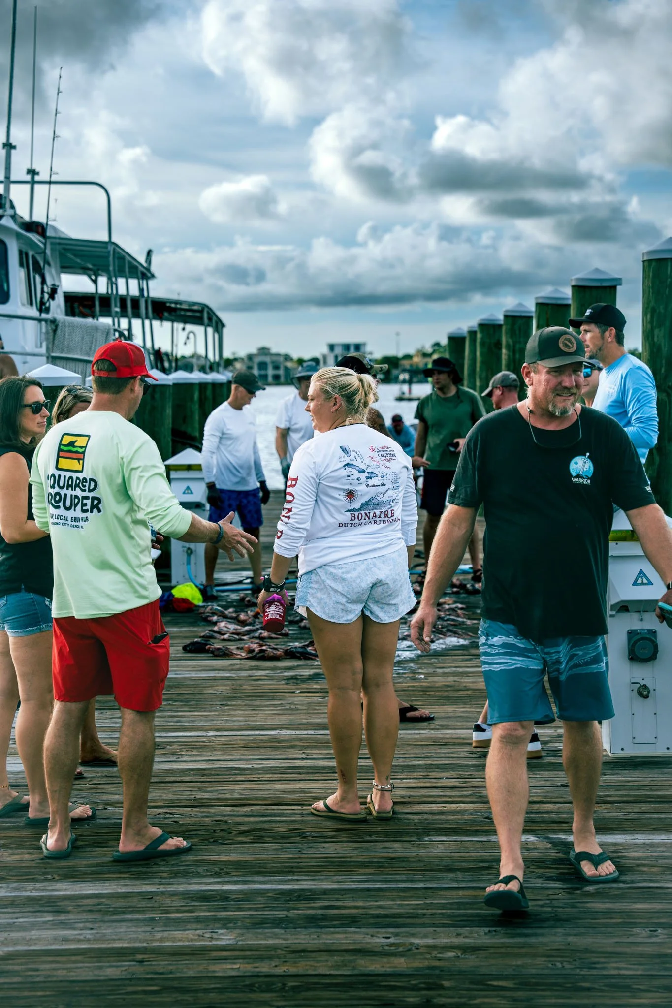 A group of people standing on a wooden dock at the Warrior Memorial Reefs Reels and Spears Fishing Tournament event at Point South Marina - Bay Point (Panama City Beach)by the water, with boats and a cloudy sky in the background.